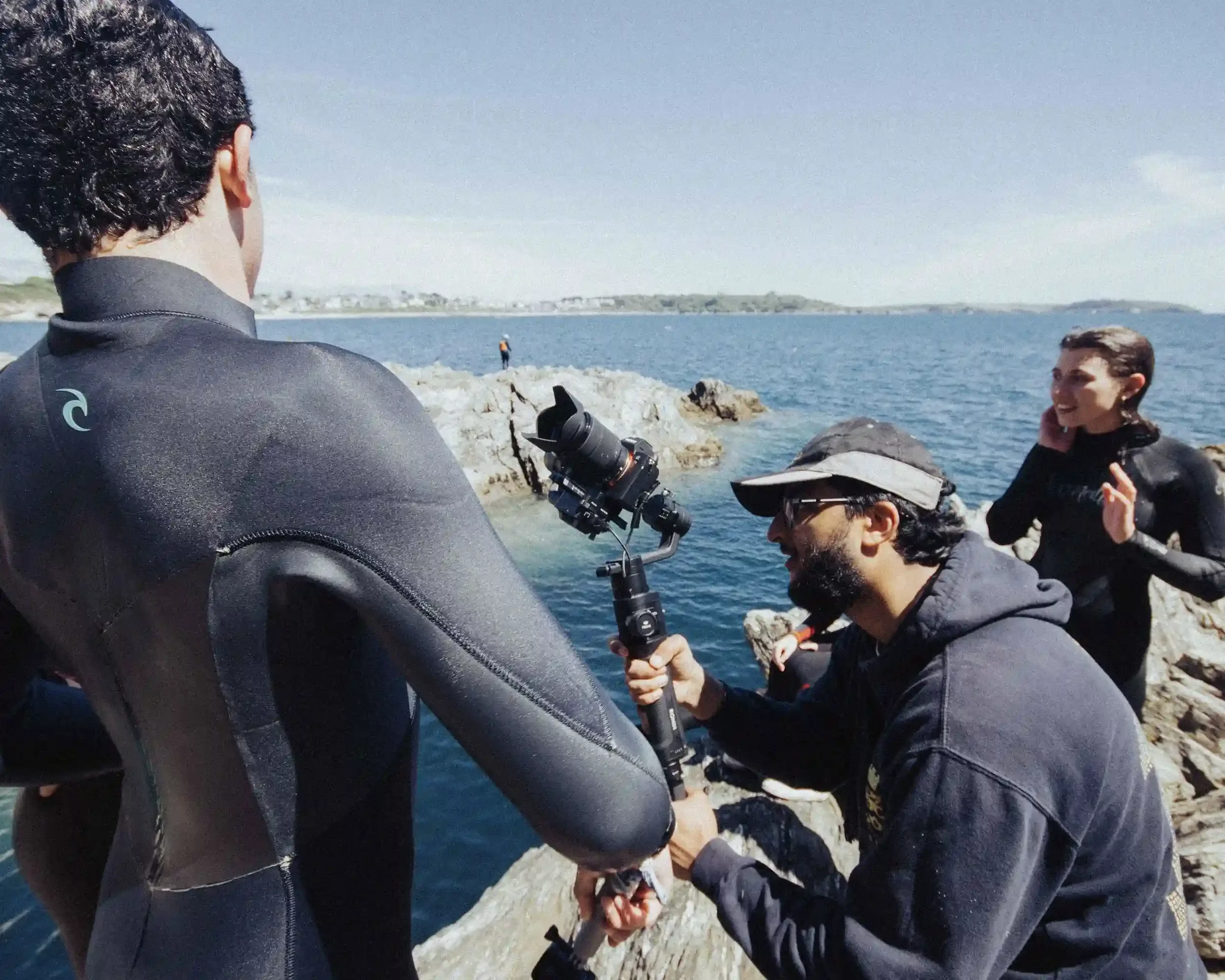 A behind-the-scenes photograph on the set of Ned Holland's 'All Things Silly' music video. A videographer in a black hoodie and cap is crouched on a rocky cliff, carefully holding a professional camera on a gimbal. In the foreground, the back of a pe