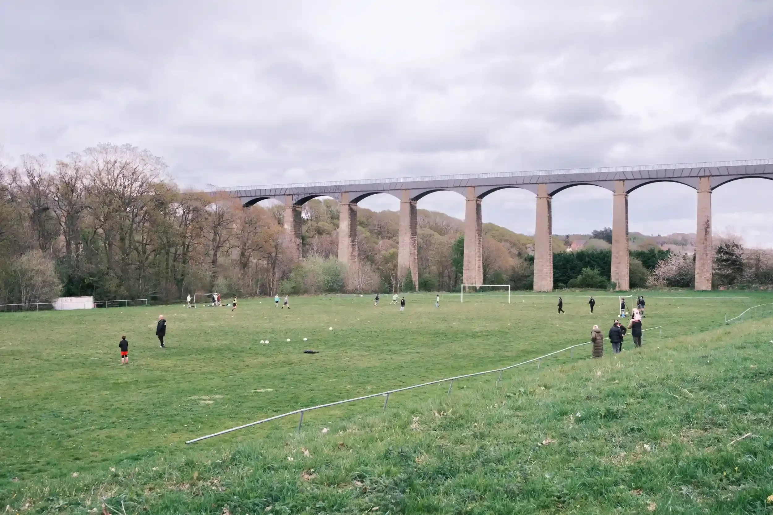 An wide-angle environmental photograph of a green football pitch where Chirk Youth FC players are training. In the background, the long, massive stone and iron structure of the Pontcysyllte Aqueduct stretches across the entire horizon against an over