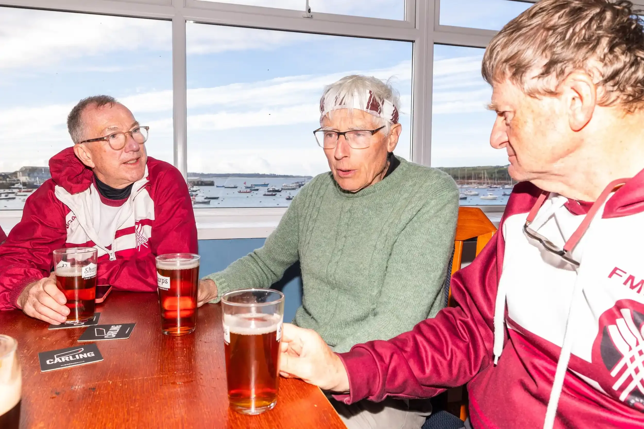 Three men from the Flushing & Mylor Pilot Gig Club sit at a wooden pub table with glasses of beer. They are wearing their team rowing gear, including maroon hoodies and technical tops. The man in the center wears a white and maroon headband. A large 