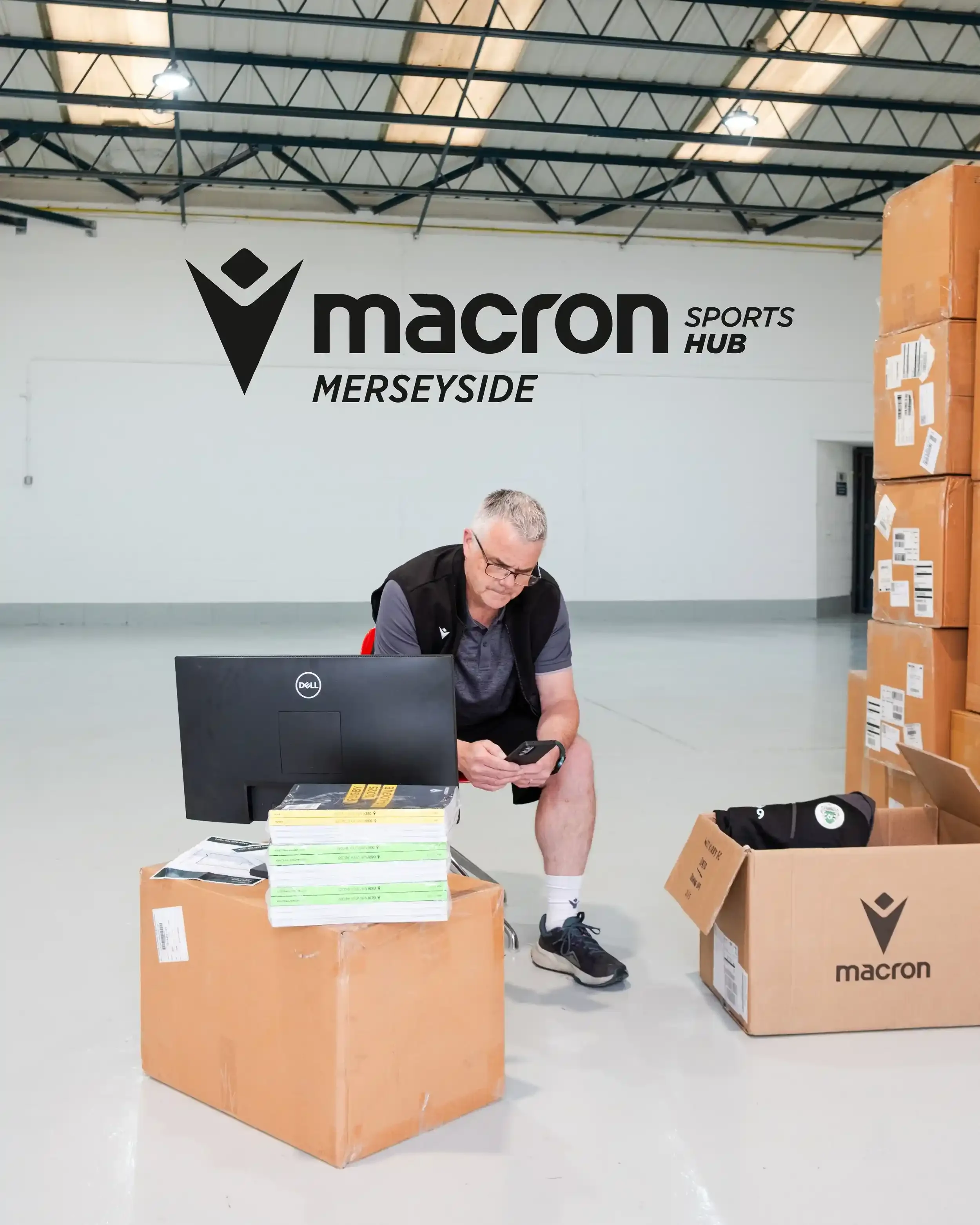 A clean, high-ceilinged warehouse interior for the launch of Macron Sports Hub Merseyside. A man in a dark grey Macron shirt and black gilet sits in the center, looking at his phone next to a Dell computer monitor. Stacks of vibrant yellow and green 