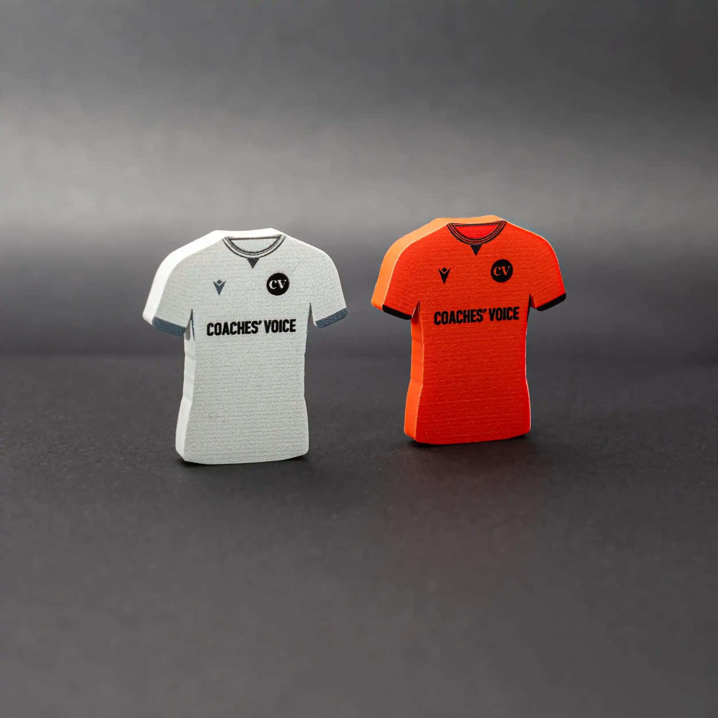 A professional studio product shot of two small, upright magnets shaped like football jerseys, standing side-by-side on a dark, textured surface. The magnet on the left is light grey with the 'Coaches' Voice' brand name and 'CV' logo. The magnet on t