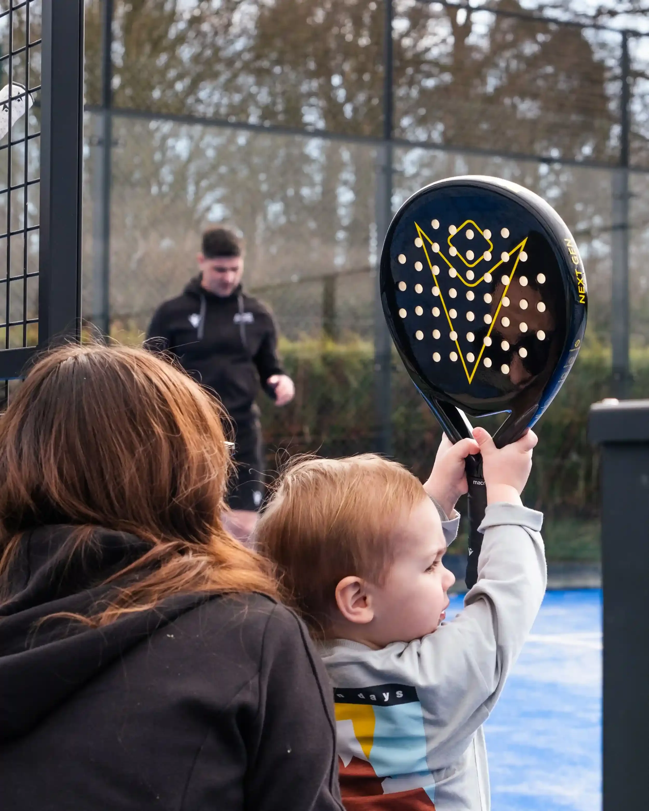 A behind-the-shoulder action shot on an outdoor padel court. A young child in a grey sweatshirt holds a glossy black Macron padel racket upright with both hands. The side of the racket features the yellow 'NEXT GEN' logo. A person with long brown hai