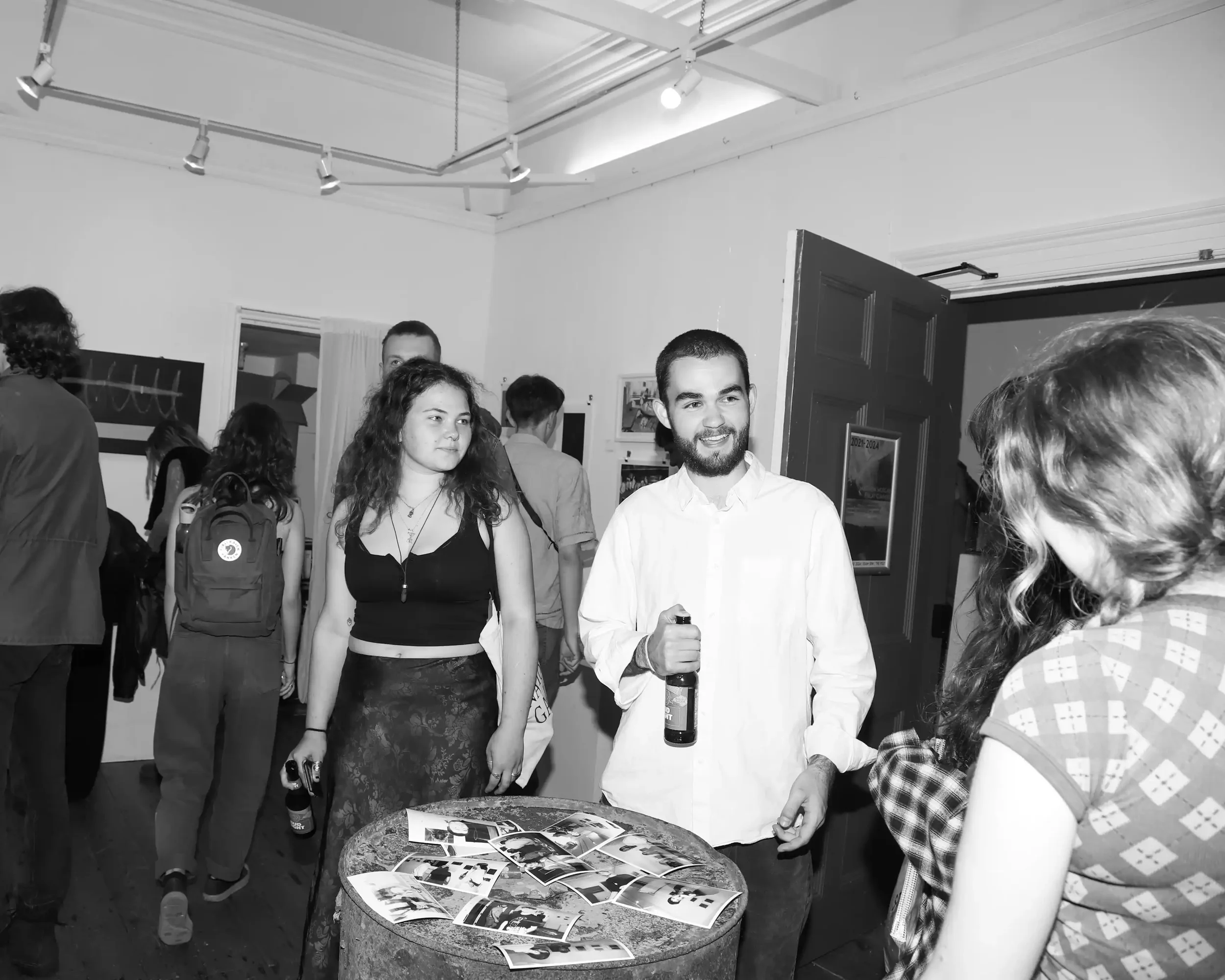 A black-and-white candid photograph of Finlay Cummins at the '2021-2024' exhibition opening night in The Poly, Falmouth. Finlay, a young man with a beard wearing a white button-down shirt, is smiling and holding a beer bottle. He stands near a rustic