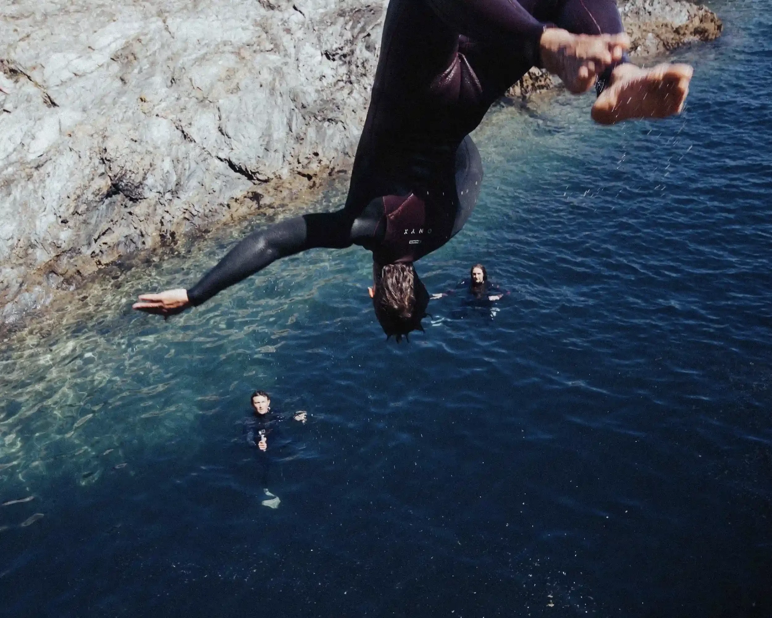 A dramatic action photograph of a person in a black wetsuit captured mid-backflip, inverted in the air above a deep blue ocean cove. Two other people are visible floating in the water below, looking up. The rugged rocky coastline and clear turquoise 