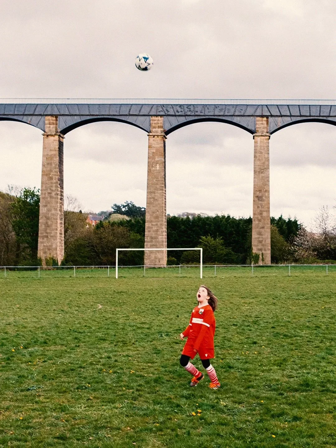 A dramatic vertical shot of a young female football player in a red Macron Chirk Youth FC kit, jumping high to head a ball. In the background, the massive stone and iron arches of the Pontcysyllte Aqueduct tower over the green pitch under an overcast