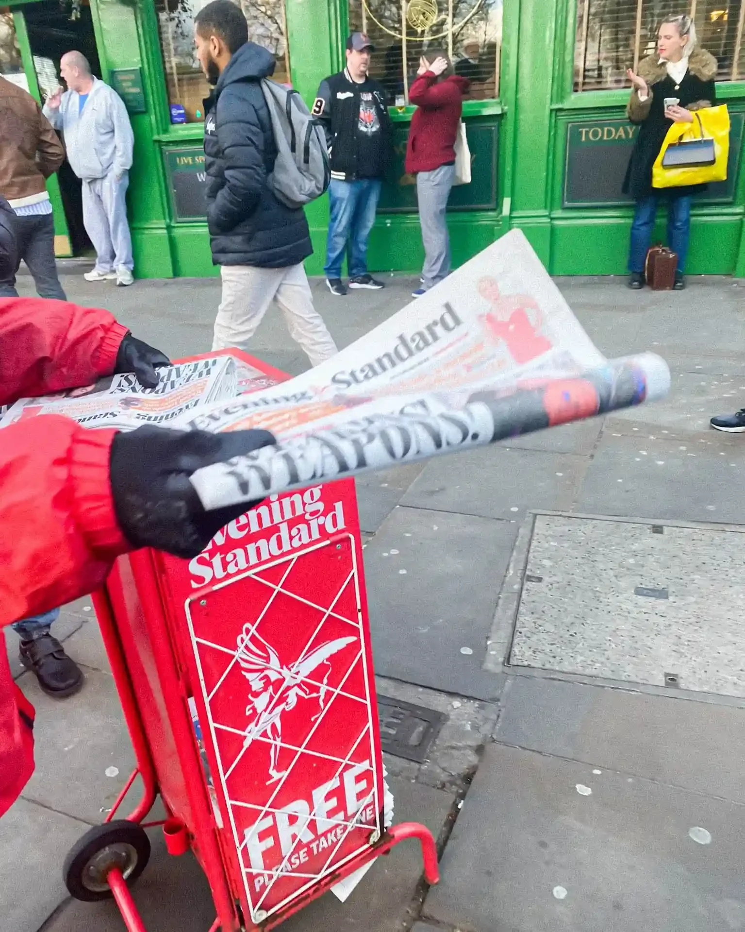 A person in a red jacket hands out free copies of the Evening Standard from a red rolling bin on a London sidewalk, captured by Matthew Morgan for his 2022 documentary series.
