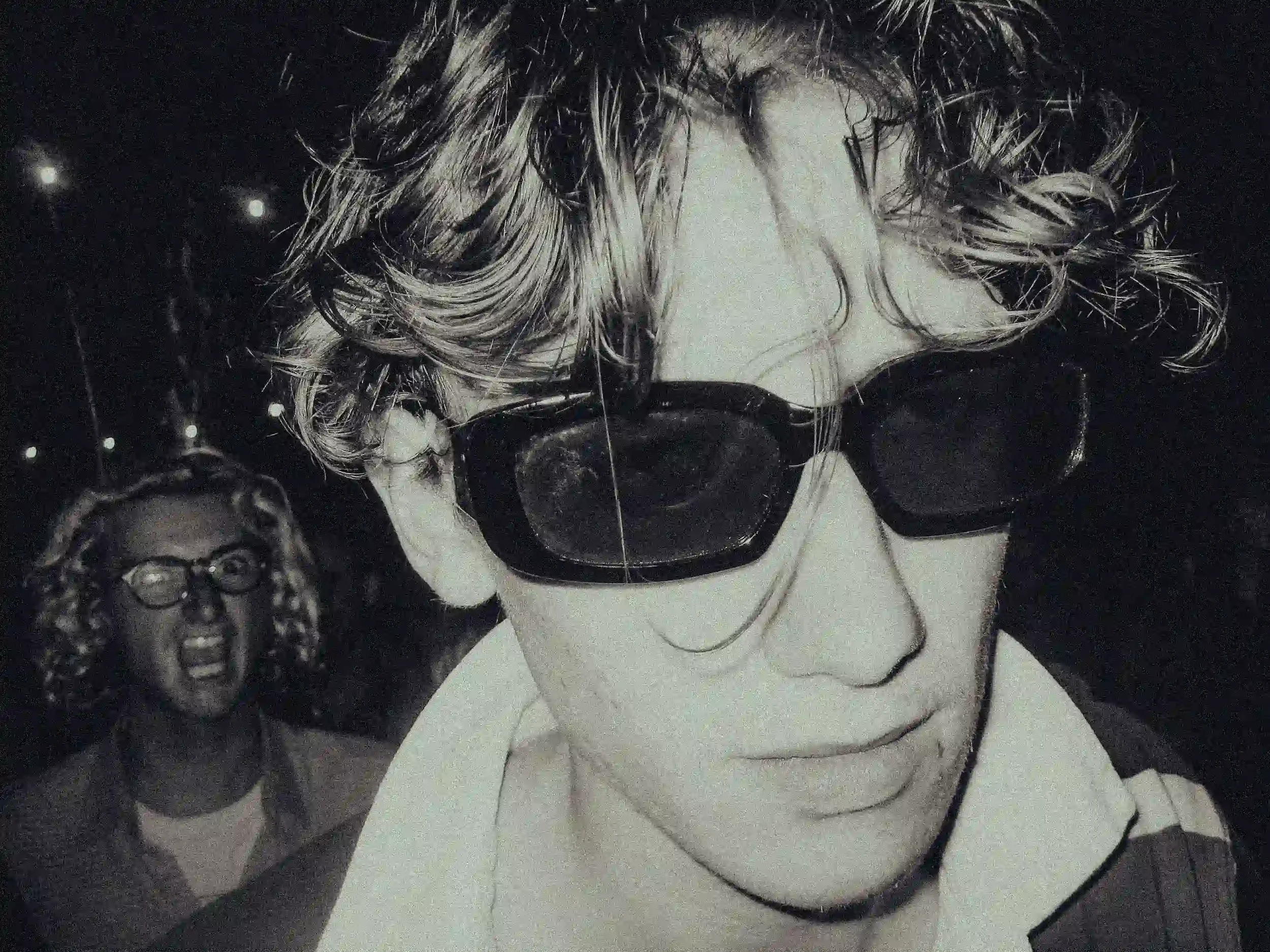 A grainy black-and-white close-up of a young man with wavy hair wearing dark rectangular sunglasses and a collared shirt. In the dark, blurred background, another person with glasses is captured mid-shout with an ecstatic expression during a performa