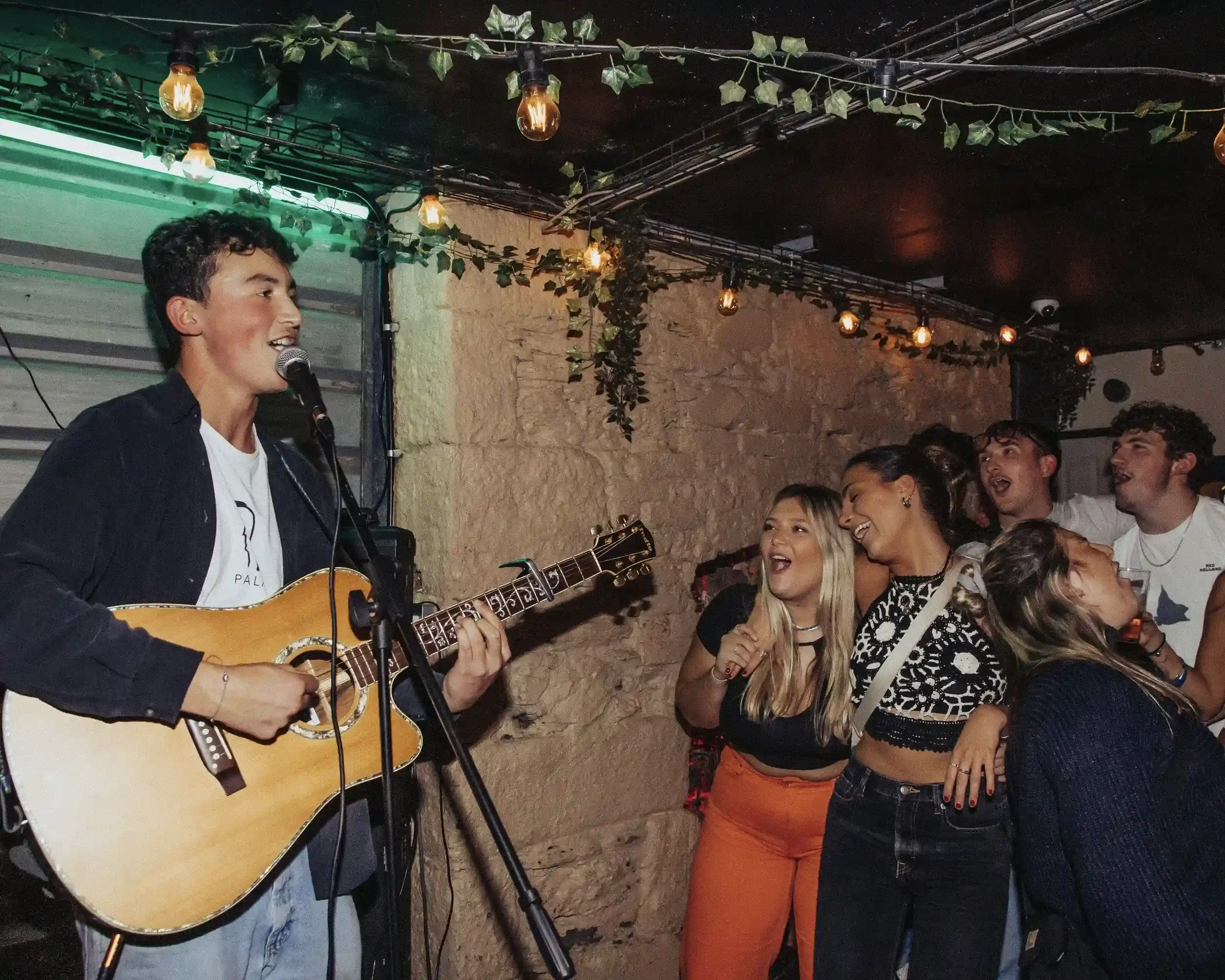A vibrant, high-contrast photograph of Ned Holland playing an acoustic guitar and singing into a microphone at Small Ships bar. He is wearing a dark jacket over a white t-shirt. To his right, a group of people are singing and laughing together agains