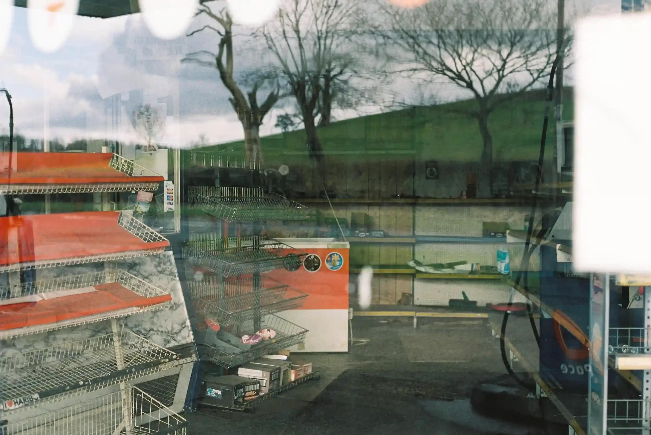 View through a window of an empty petrol station shop in Tewkesbury with bare red and white metal shelves and a reflection of bare trees and a green hill in the glass, by Matthew Morgan.