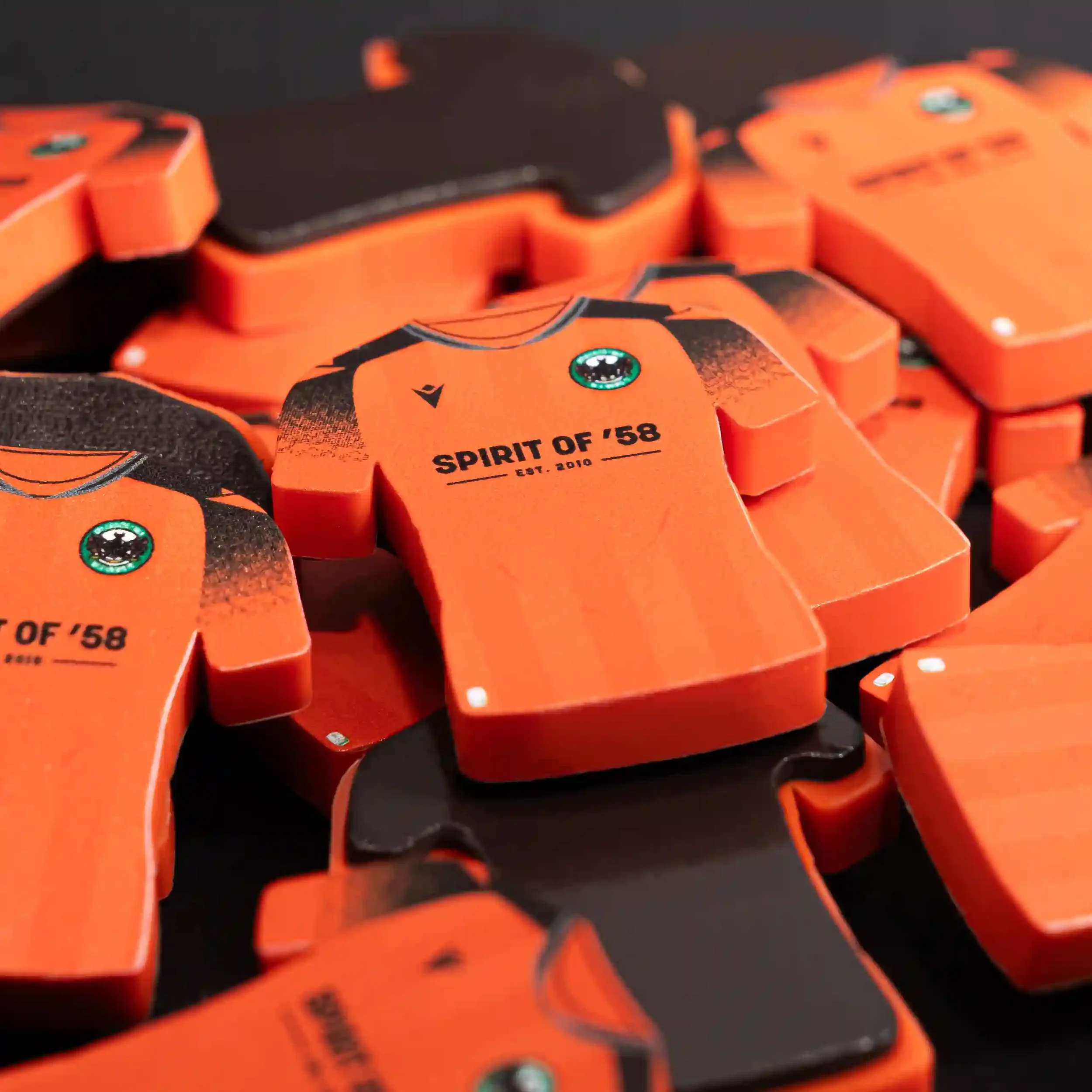 A close-up product photograph of several small, shirt-shaped commemorative items in bright orange with black accents. The items feature the Bala Town FC club crest and the text 'SPIRIT OF '58 EST. 2010' printed on the chest. The products are piled to