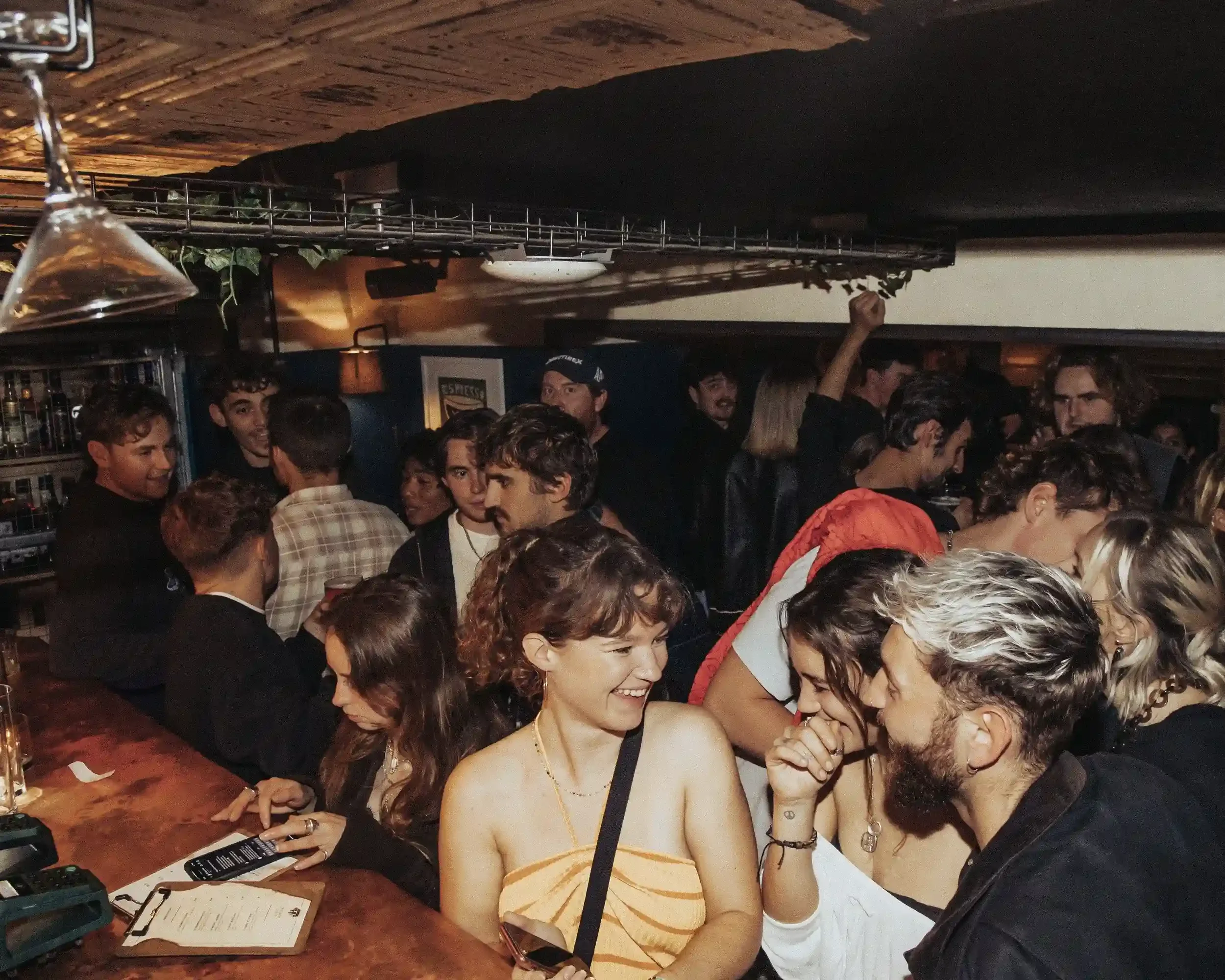 A vibrant, crowded scene at the bar inside Small Ships. In the foreground, a woman in a yellow top and a man with bleached hair are laughing together. The bar is packed with people talking and ordering drinks under a low, dark ceiling. The direct fla
