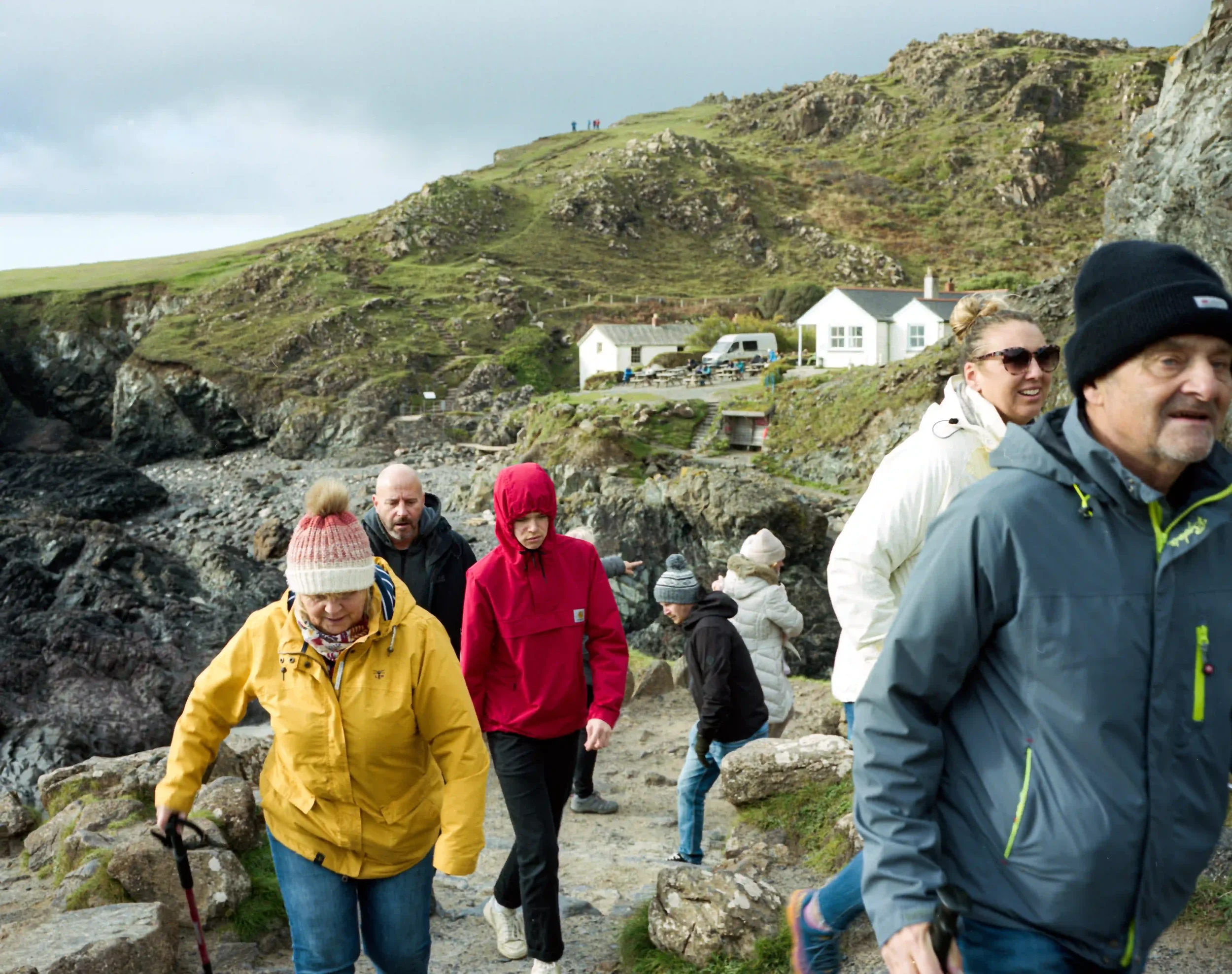 A dynamic, medium-wide shot of several people in colorful winter gear, including a yellow raincoat, a red hooded jacket, and a blue parka, walking up a rough stone path at Kynance Cove. In the background, white-washed coastal buildings sit at the bas