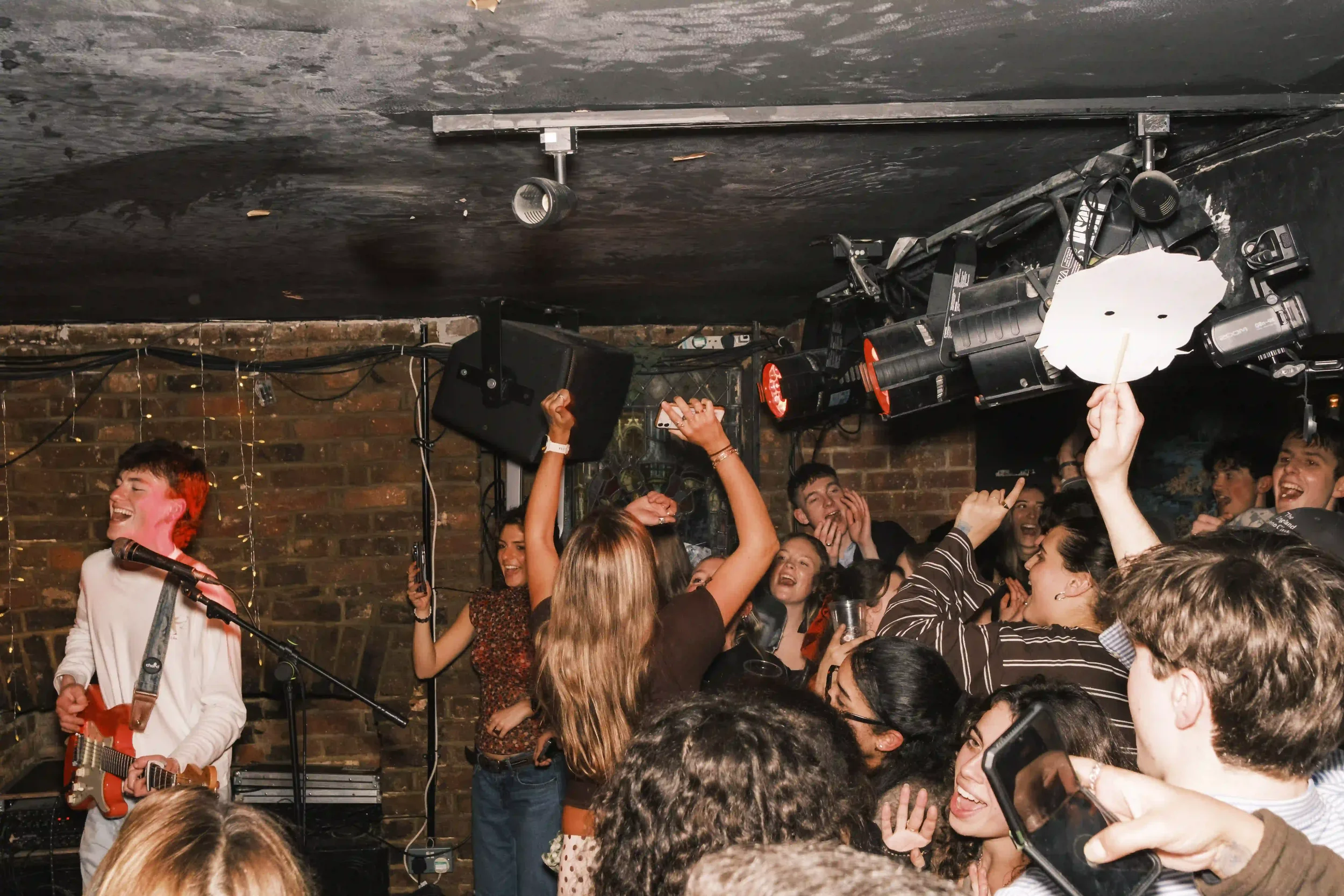 A wide-angle live music photograph of Tom Scamell performing at The Troubadour, London. On the left, Scamell is singing and playing a red electric guitar, illuminated by warm stage lights against a brick wall. In the foreground and center, a dense cr