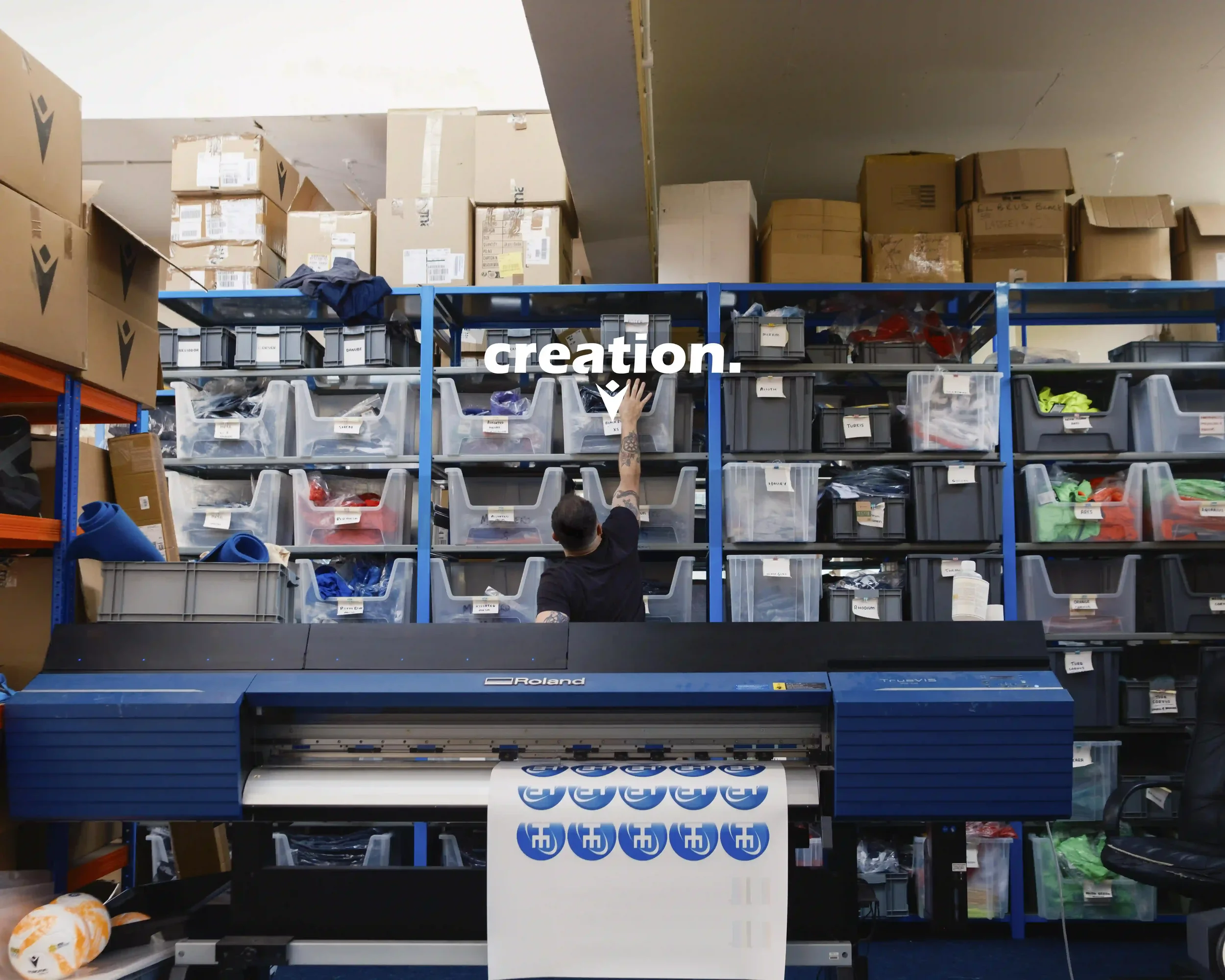 A wide-angle shot of the industrial printing and storage room at Macron Sports Hub Wrexham. In the foreground, a blue Roland TrueVIS printer is outputting a sheet of circular blue and white club logos. In the background, a man in a black t-shirt reac