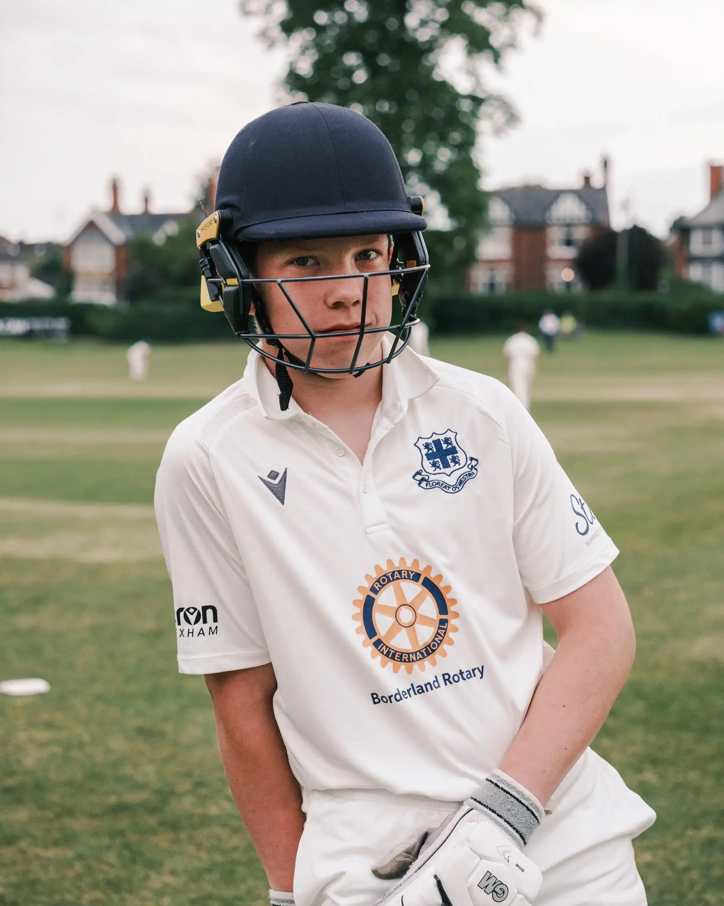 A focused portrait of a young cricketer from Oswestry CC on a green field. He is wearing a navy blue batting helmet and a white Macron cricket shirt with the club crest on the left chest and the Borderland Rotary logo in the center. The background sh