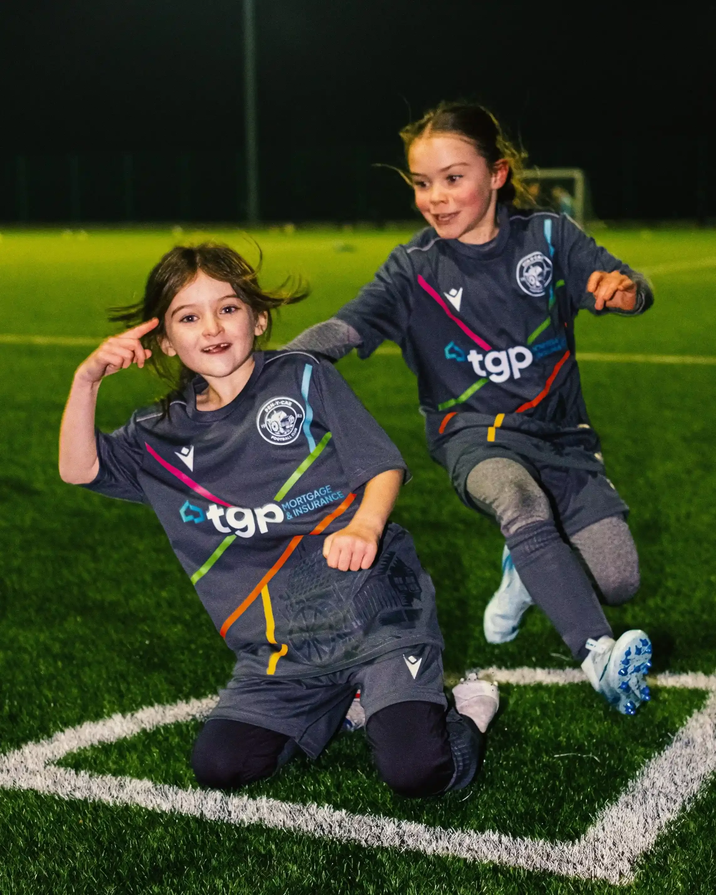A dynamic, low-angle action shot of Tess, a young girl, performing a celebratory knee slide on a green artificial turf pitch at night. She is smiling and pointing to her head with one hand, wearing the custom grey Macron kit she designed, which featu