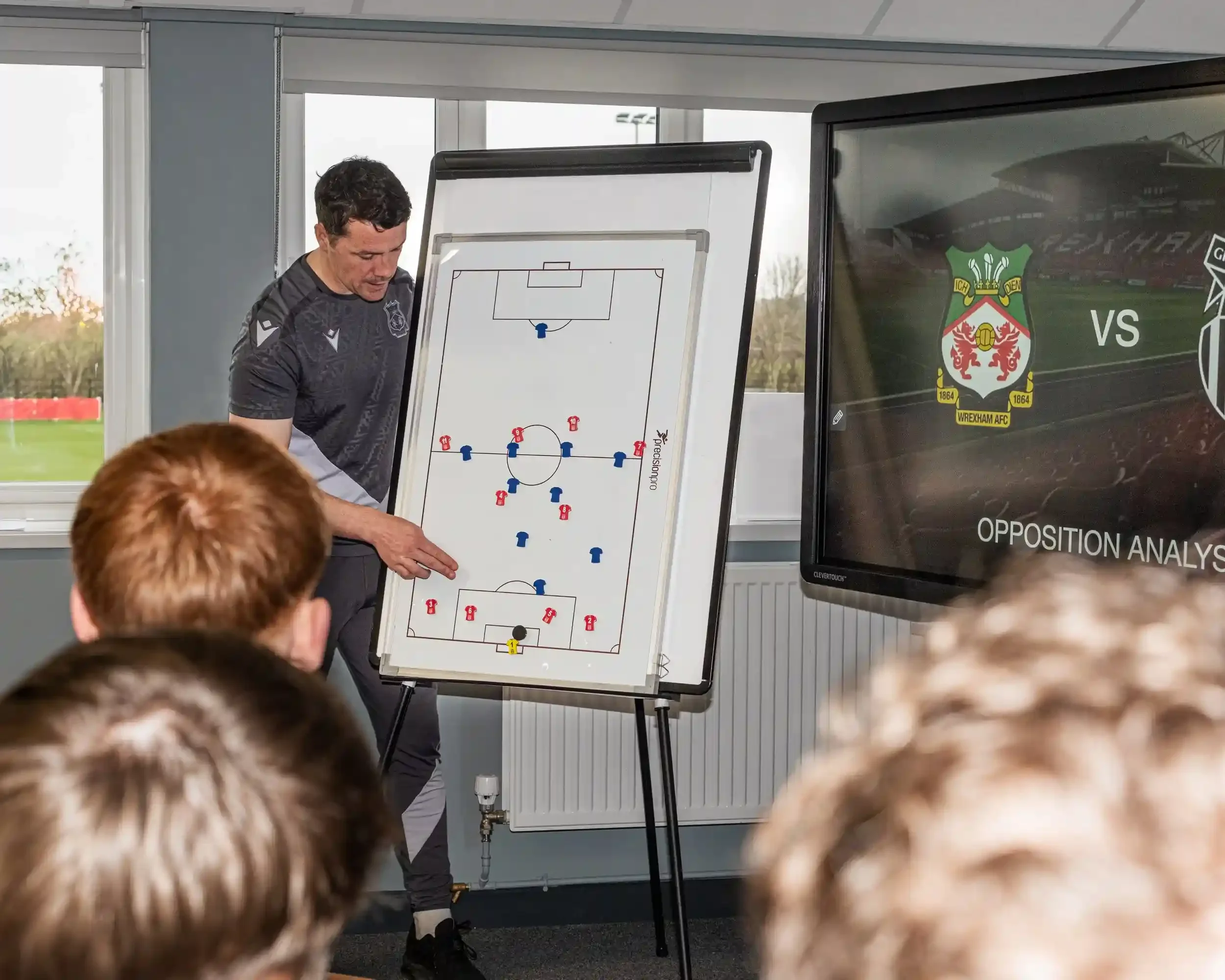 A wide shot of a tactical briefing room at the FAW training ground. A coach in a grey Wrexham AFC training kit stands between a physical whiteboard and a large digital monitor. He is pointing to custom red and blue tactical magnets from Your Game You