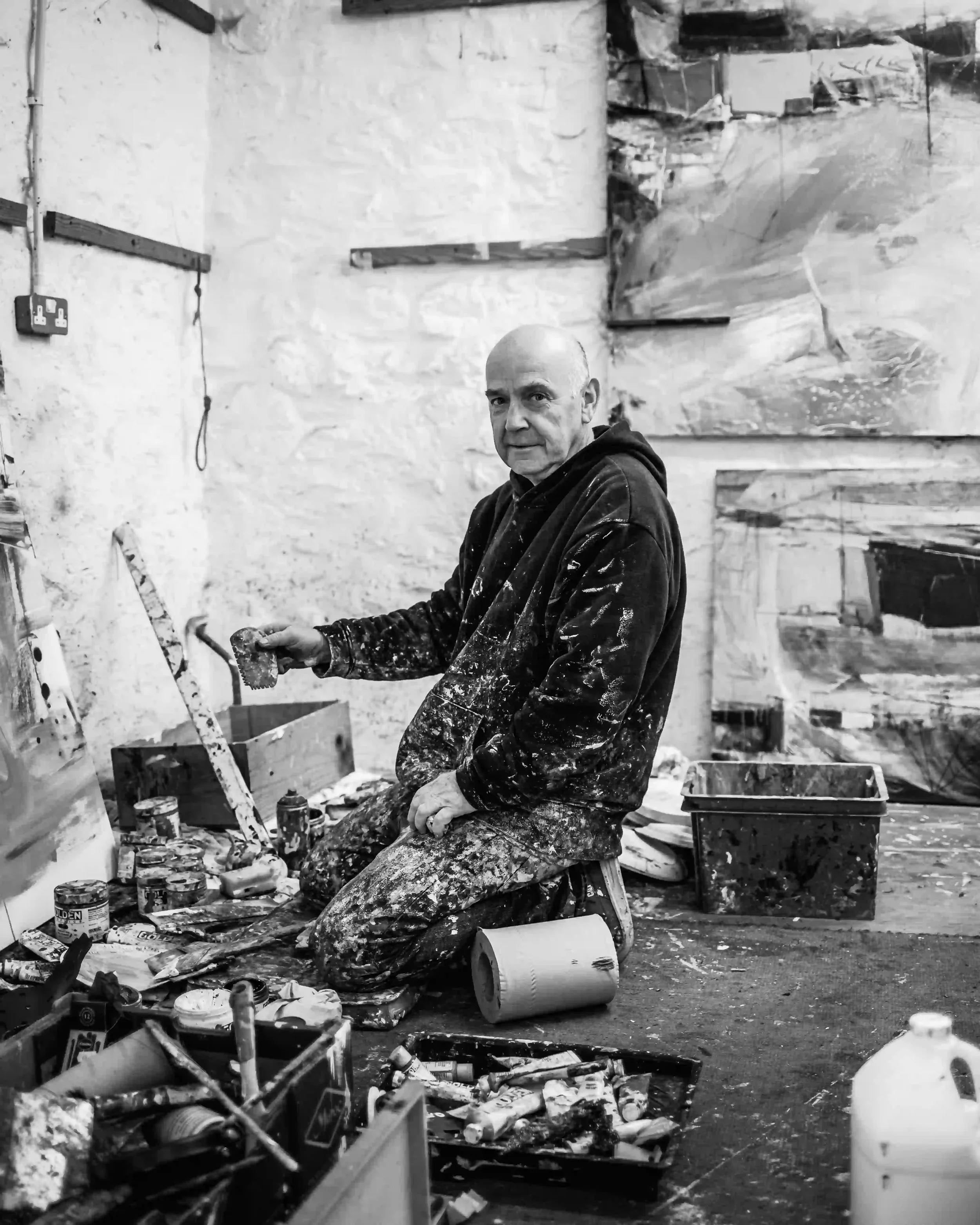 A wide-angle editorial shot of artist David Mankin in his light-filled Cornwall studio, surrounded by large-scale abstract canvases. He is captured in a moment of 'excavation,' working with mixed media and acrylics to create the layered textures and 