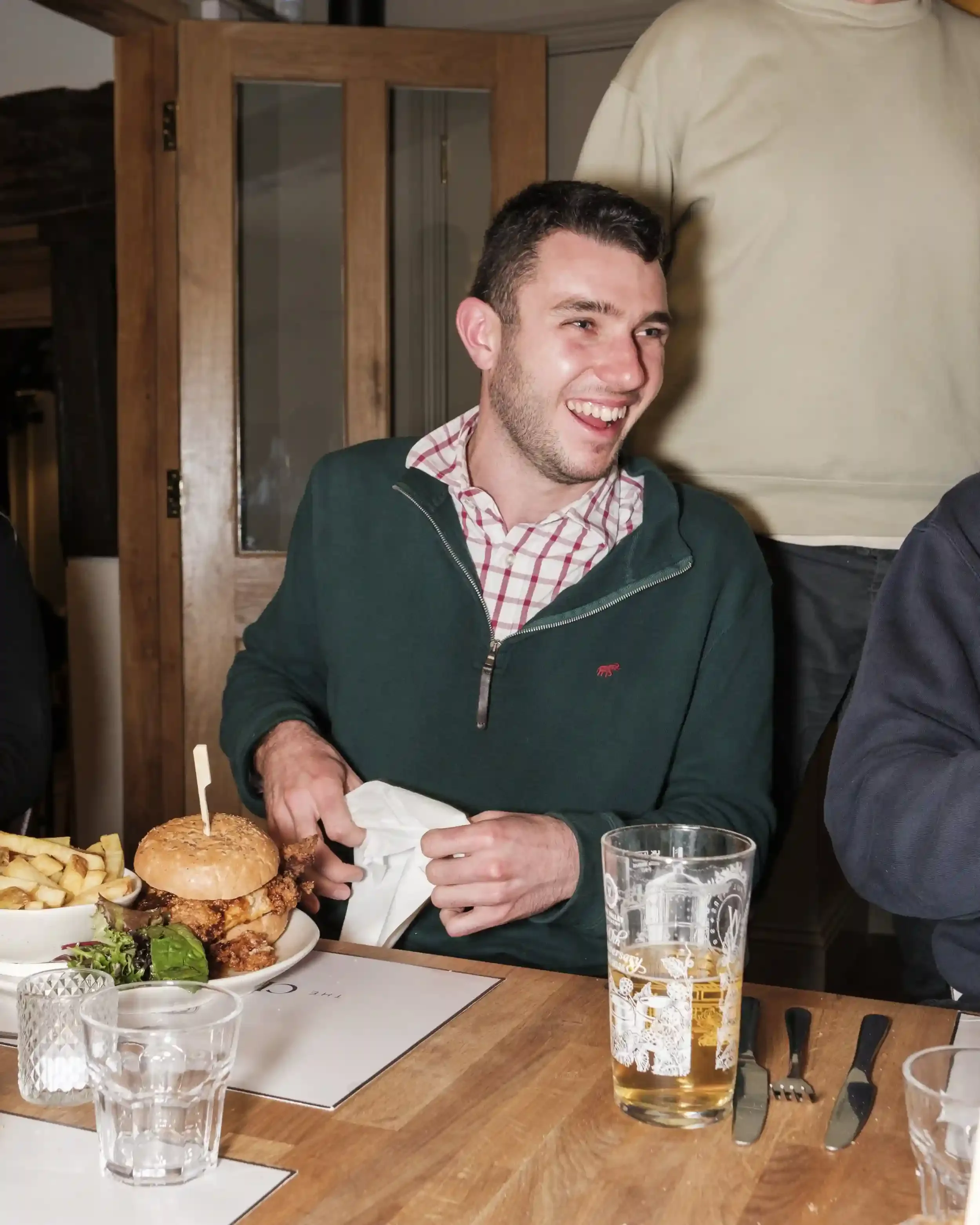 A candid photograph by Matthew Morgan of a man laughing while seated at a wooden dining table at The Crown Inn, Lea. He is wearing a green quarter-zip sweater over a red and white checkered shirt. In front of him is a large gourmet chicken burger in 