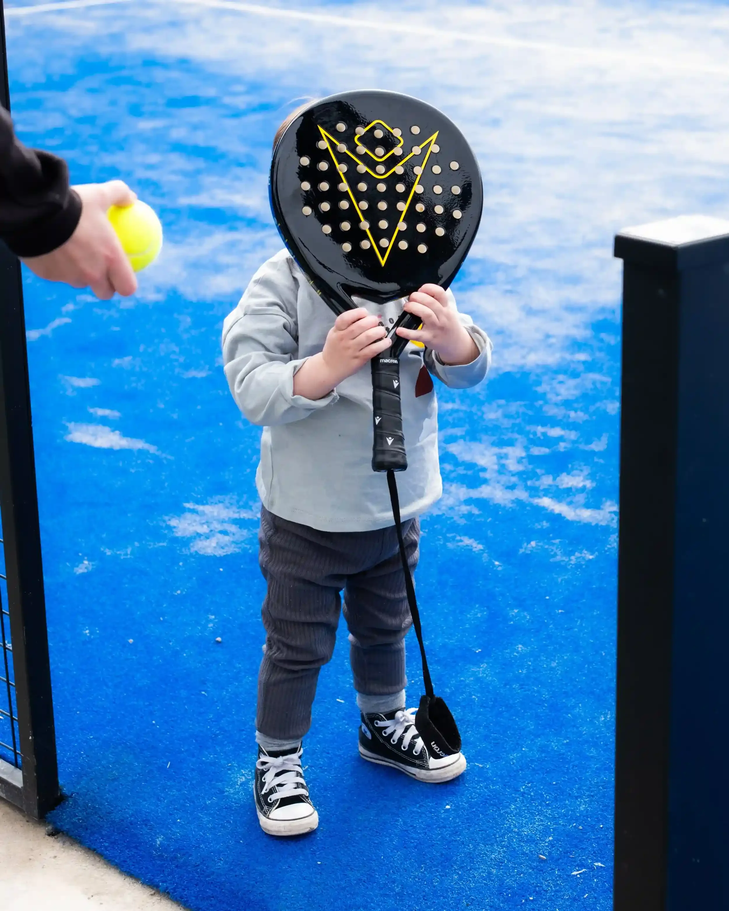 A vertical lifestyle shot on a bright blue padel court. A small child in a grey sweatshirt and dark trousers holds a glossy black Macron padel racket vertically, obscuring their face. The racket features a prominent yellow 'M' logo and a perforated s
