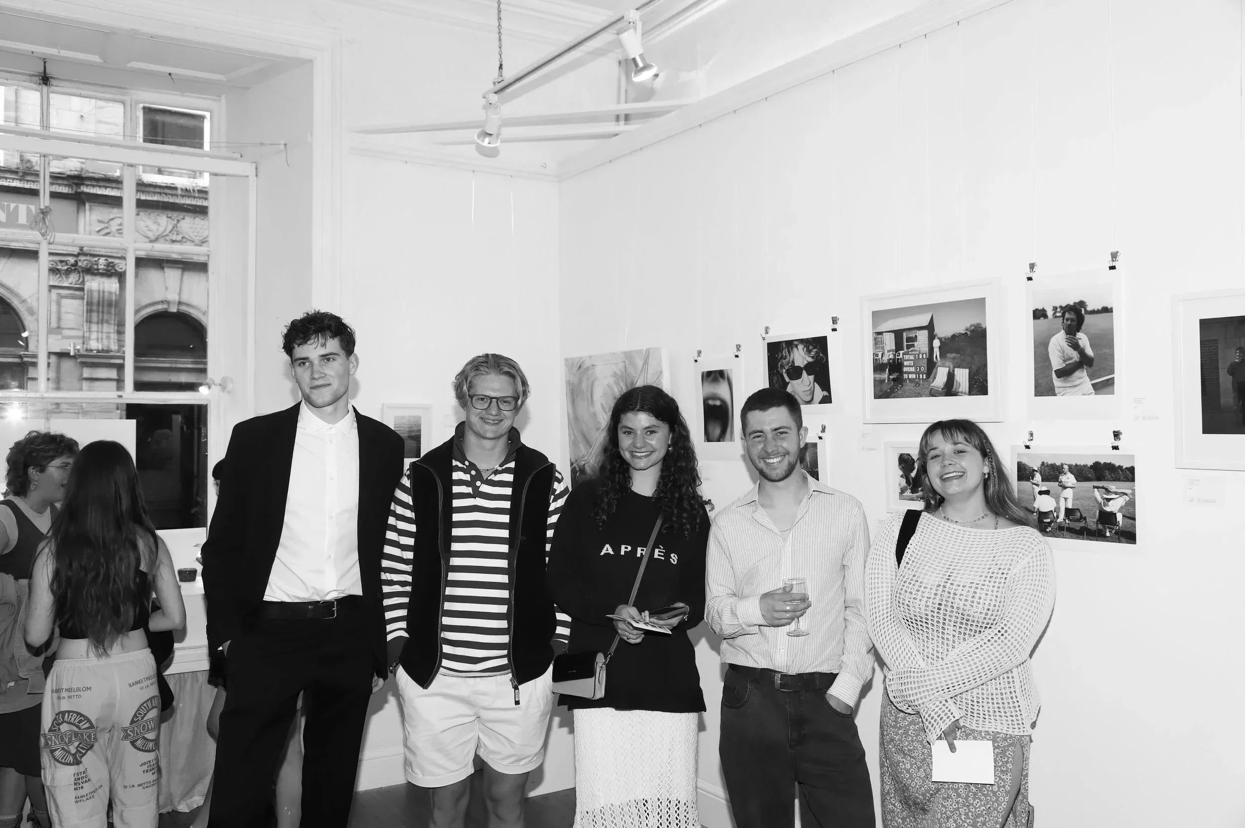 A black-and-white group portrait taken during the '2021-2024' exhibition at The Poly in Falmouth. Five young people, including the artist Matthew Morgan, are standing together and smiling in front of a white gallery wall. The wall is decorated with a