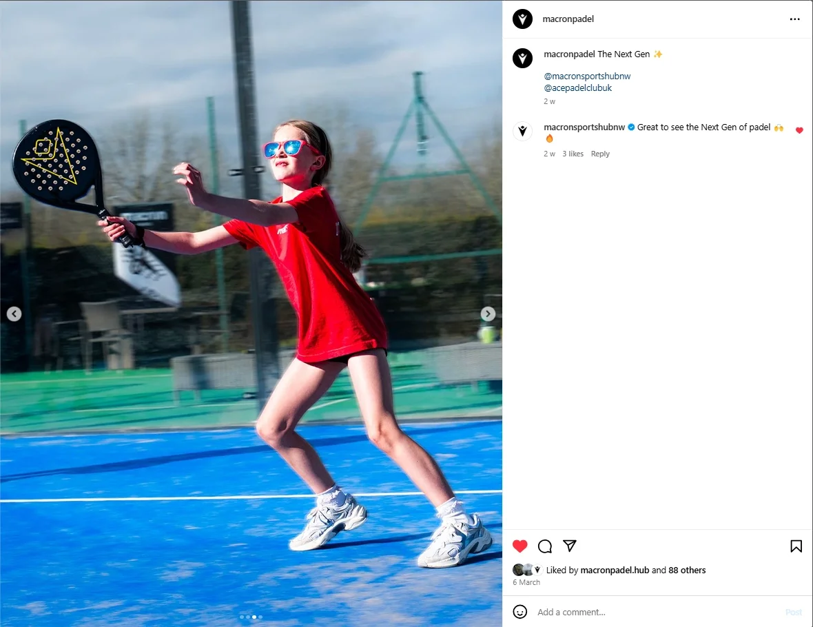 A dynamic outdoor action shot of a young girl playing padel on a vibrant blue court. She is wearing a red shirt and bright pink sunglasses, lunging forward while holding a black Macron padel racket with yellow branding. The background is a crisp, bri