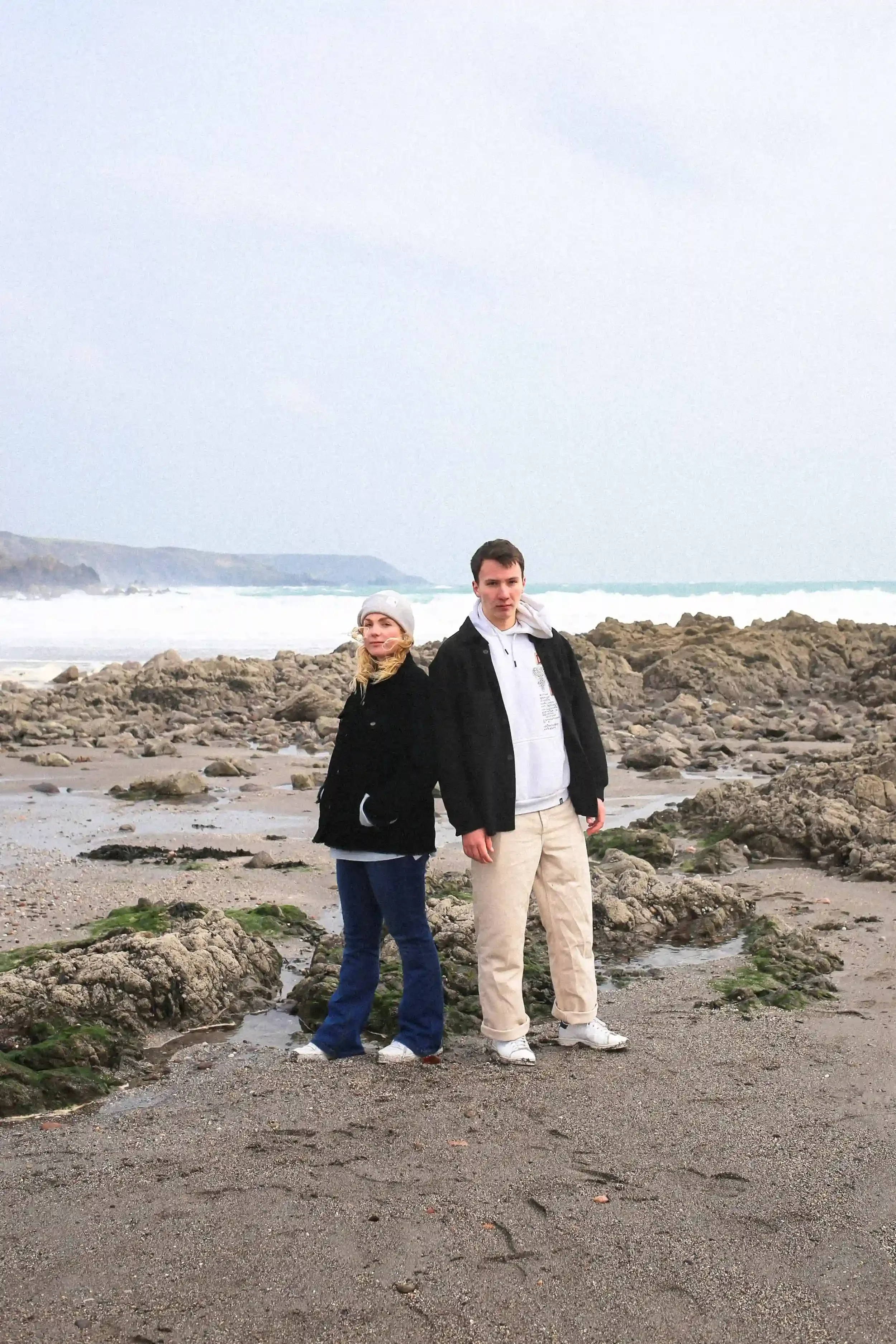 A colour film unit still portrait of a woman in a black jacket and grey beanie and a man in a black jacket and white hoodie standing on a rugged, rocky beach. They are standing back-to-back, looking directly into the lens. The background features whi