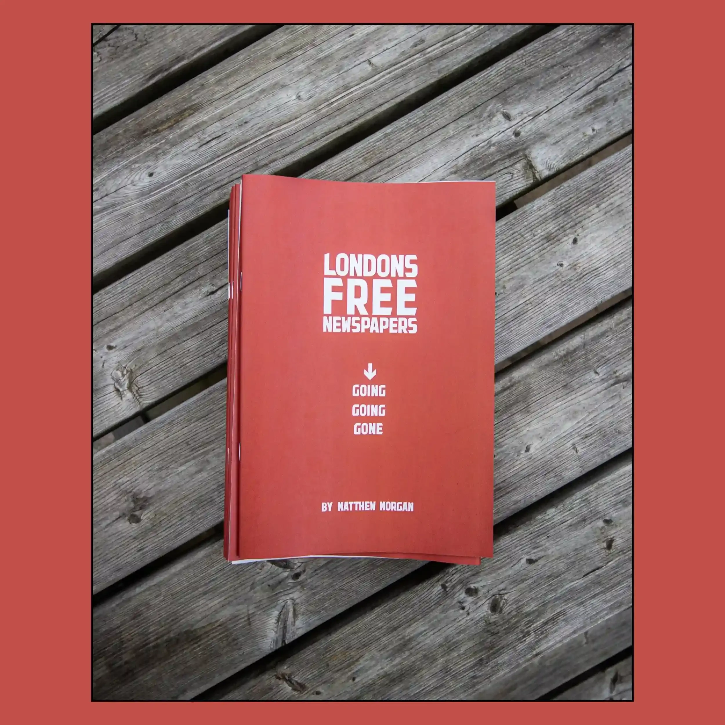 The red cover of Matthew Morgan's photography zine titled 'London's Free Newspapers: Going, Going, Gone' resting on a wooden slatted surface.
