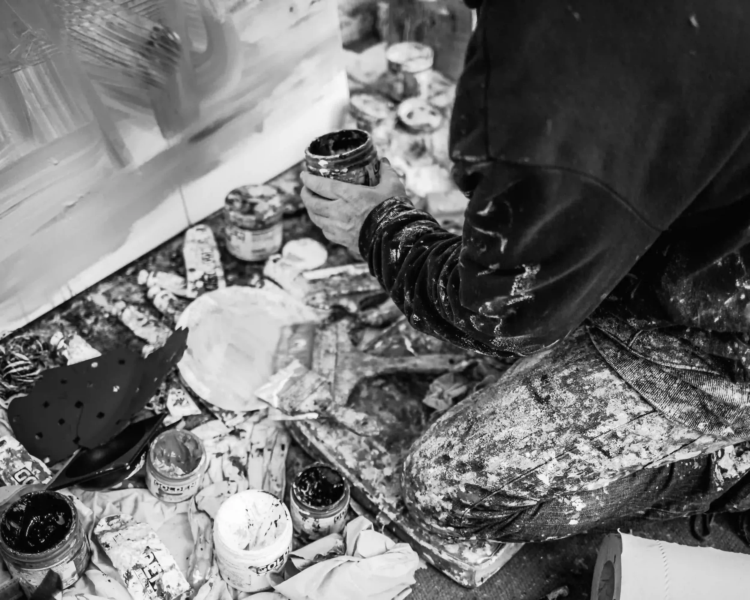 A high-angle, black-and-white close-up of an artist's paint-splattered trousers and hands as they work on the studio floor. The person is holding a small jar of paint, surrounded by an array of open paint containers, messy brushes, and used sandpaper