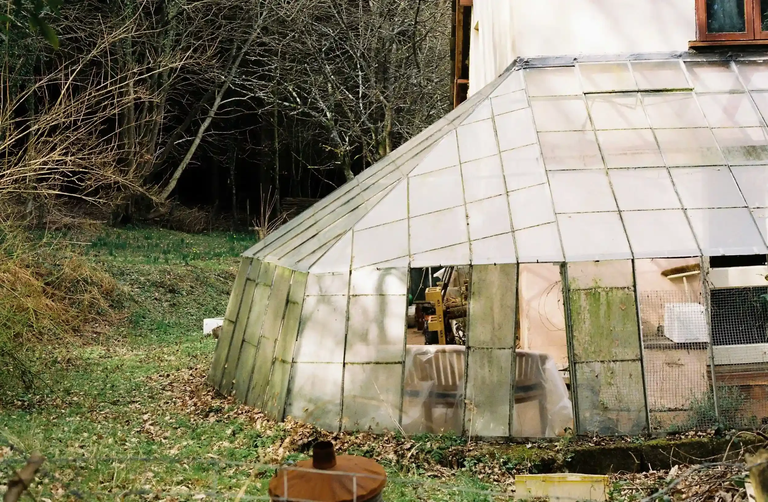 A large, angled glass greenhouse with weathered, mossy panes sitting at the edge of a wooded area near May Hill, with a wooden chair visible through the glass, by Matthew Morgan.
