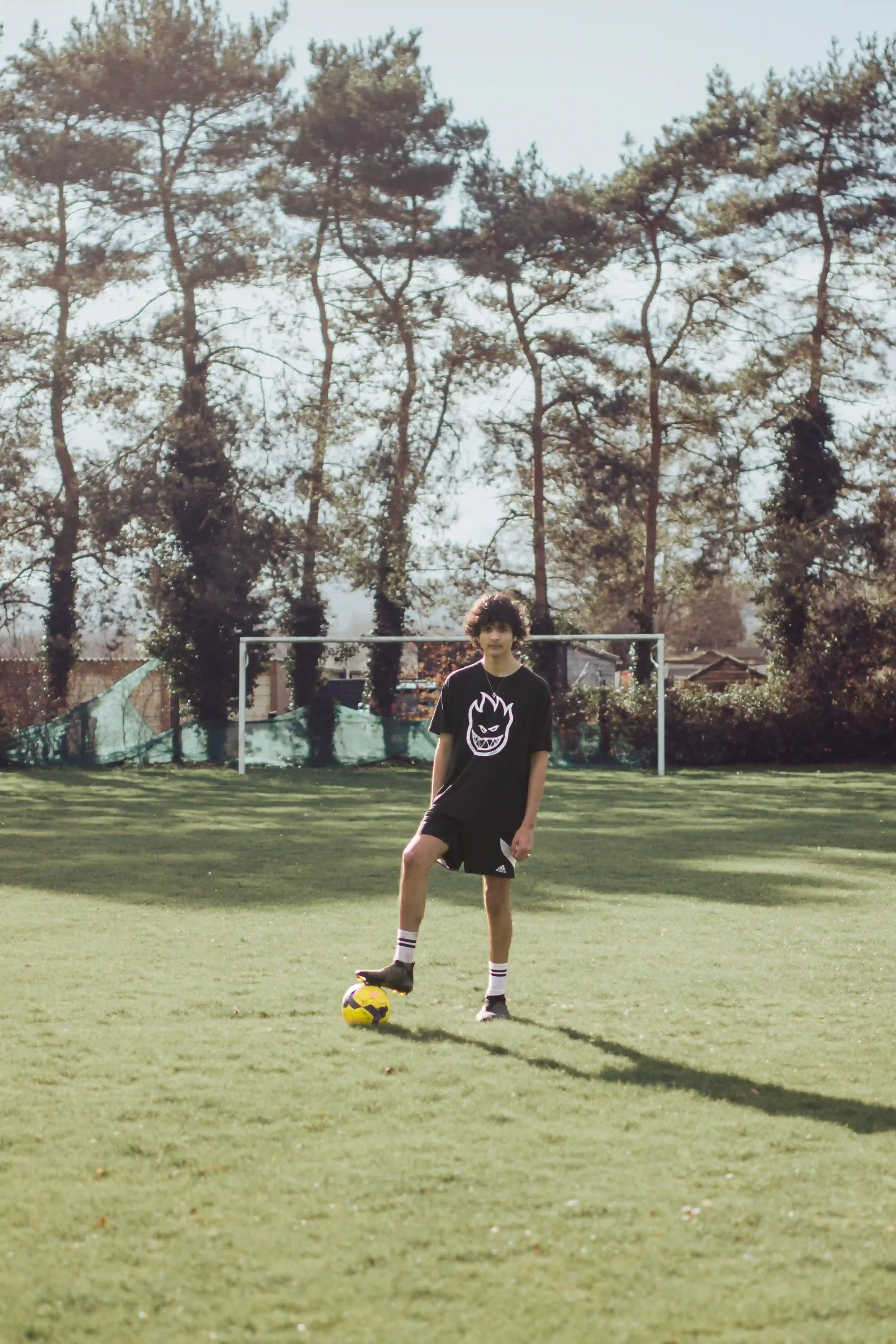 A young man, Mason Townsend, stands on a sunlit green grass football pitch with one foot on a yellow soccer ball. He is wearing a black Spitfire t-shirt and shorts, with a white goalpost and a line of tall pine trees in the soft-focus background, by 