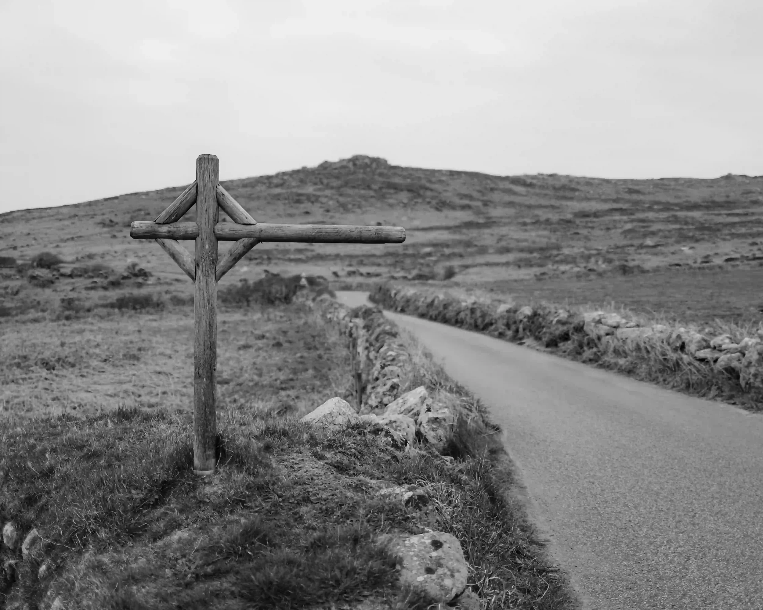 A black-and-white photograph of a simple, cross-shaped wooden signpost without text, standing in the grassy verge of a narrow, winding road near St Just. Low stone walls line the road as it leads toward a misty hill or tor in the distance, by Matthew