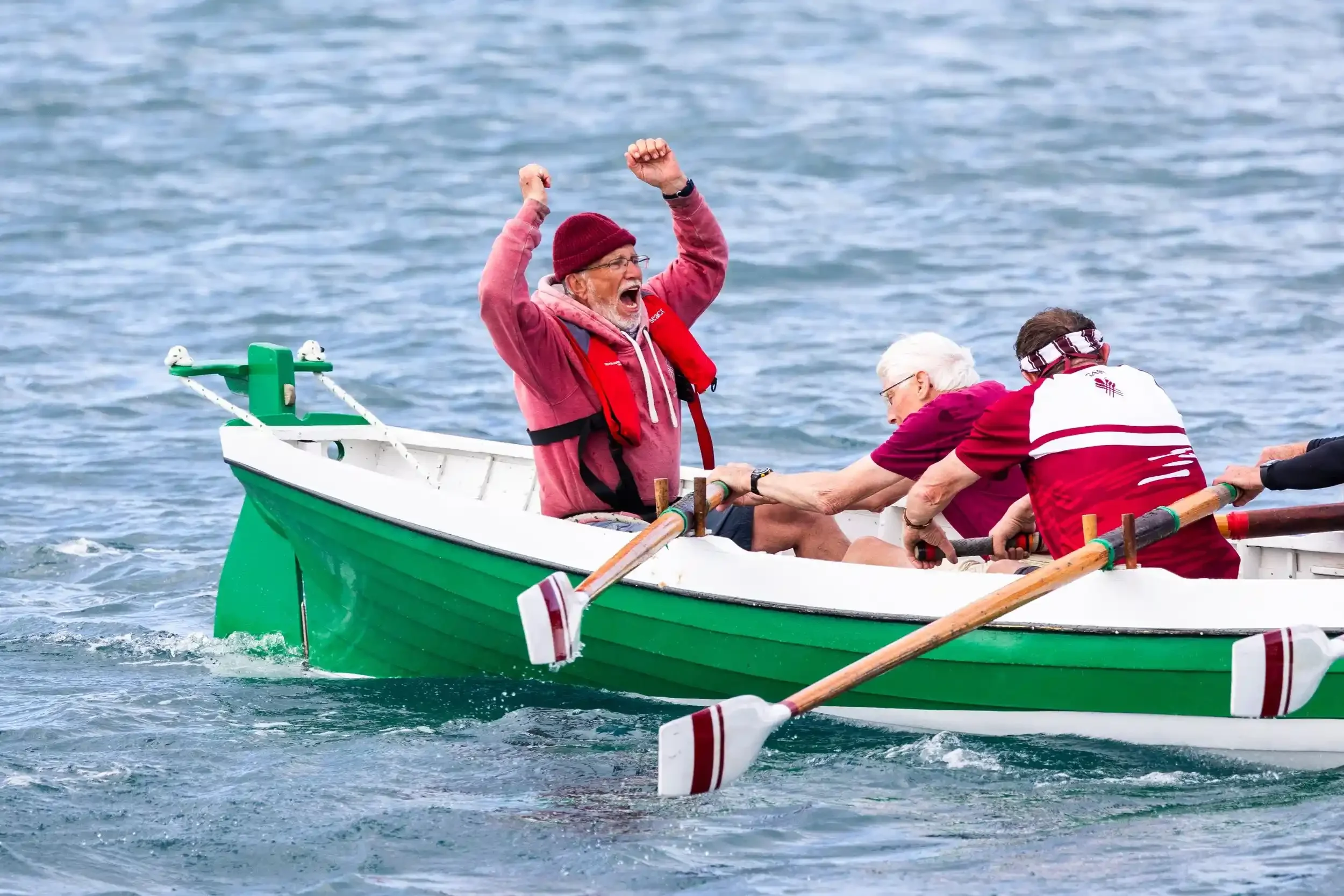 A dynamic sports photograph of a rowing crew in a green and white wooden pilot gig. In the stern, the coxswain, wearing a red beanie and life jacket, has both fists raised in the air with his mouth open in an encouraging shout. The rowers in front of