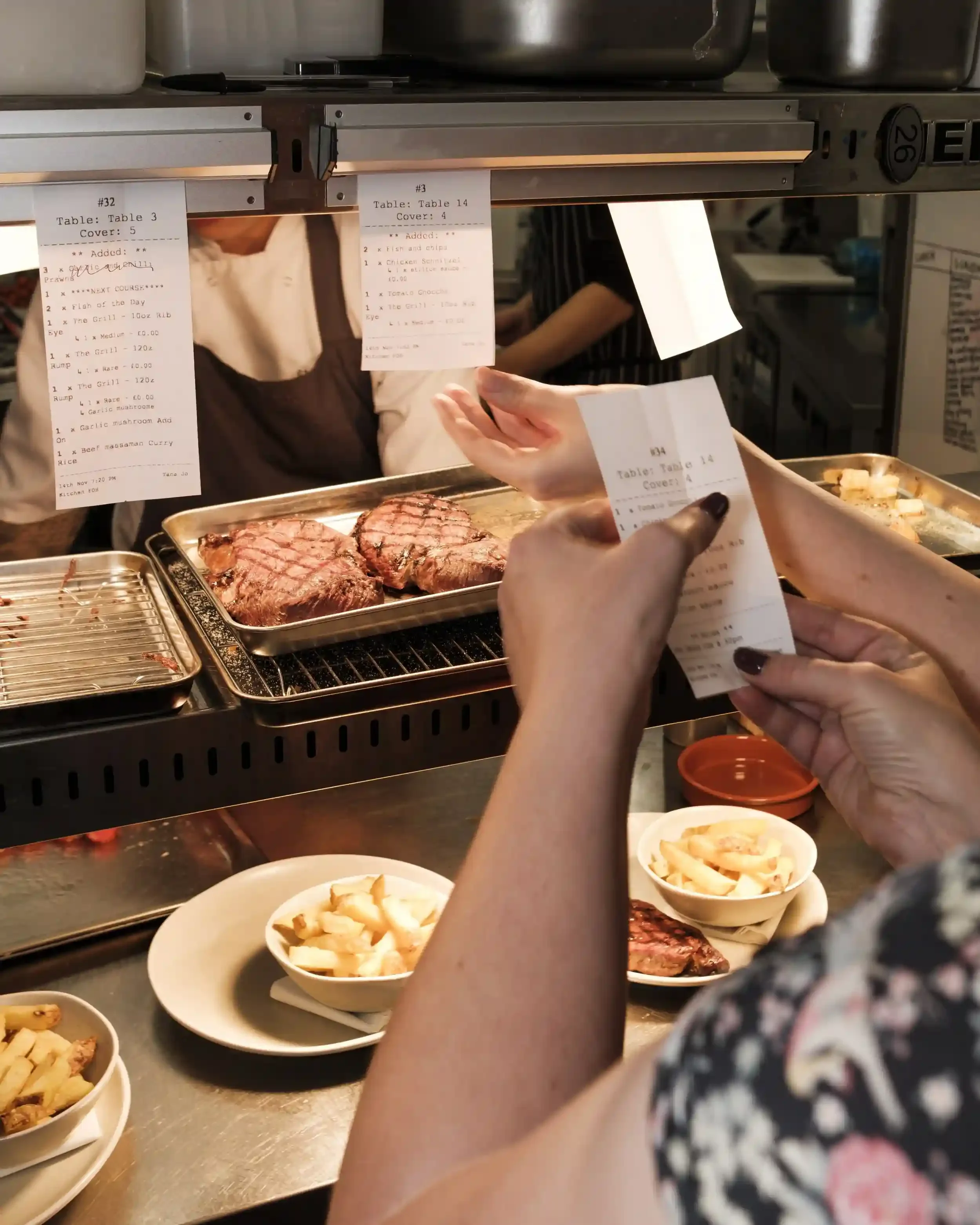 A busy kitchen scene at The Crown Inn, Lea, photographed by Matthew Morgan for SoSocial Media. The foreground shows a person holding a kitchen order ticket for 'Table 14,' listing a 'Chicken Schnitzel' and 'Tomato Gnocchi.' On the metal pass, two per