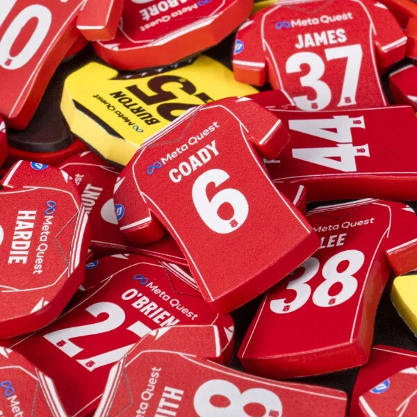 A top-down, close-up photograph of several red and yellow Wrexham AFC tactical magnets piled together. The red magnets are shaped like the club's home jersey and feature white text for various player names and numbers, including 'COADY 6', 'O'BRIEN 2