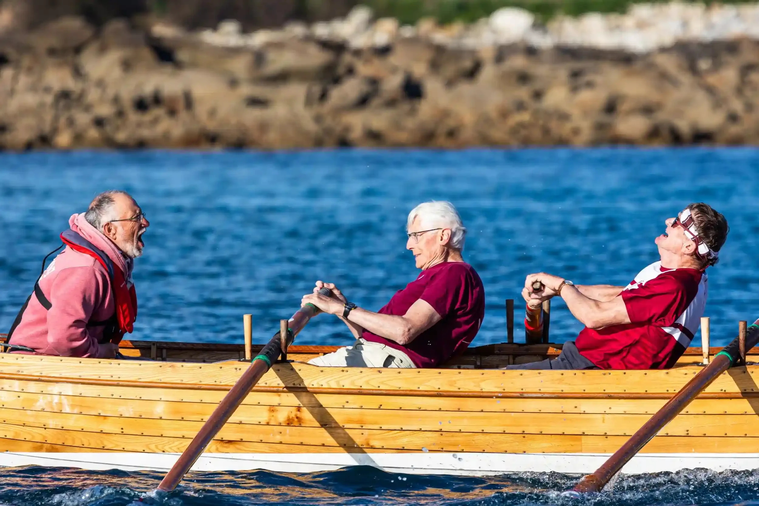 A close-up action shot of three men in a traditional wooden pilot gig rowing through bright blue sea water. The man on the left, wearing a pink hoodie and life jacket, has his mouth open as if shouting encouragement. The two rowers in the middle and 