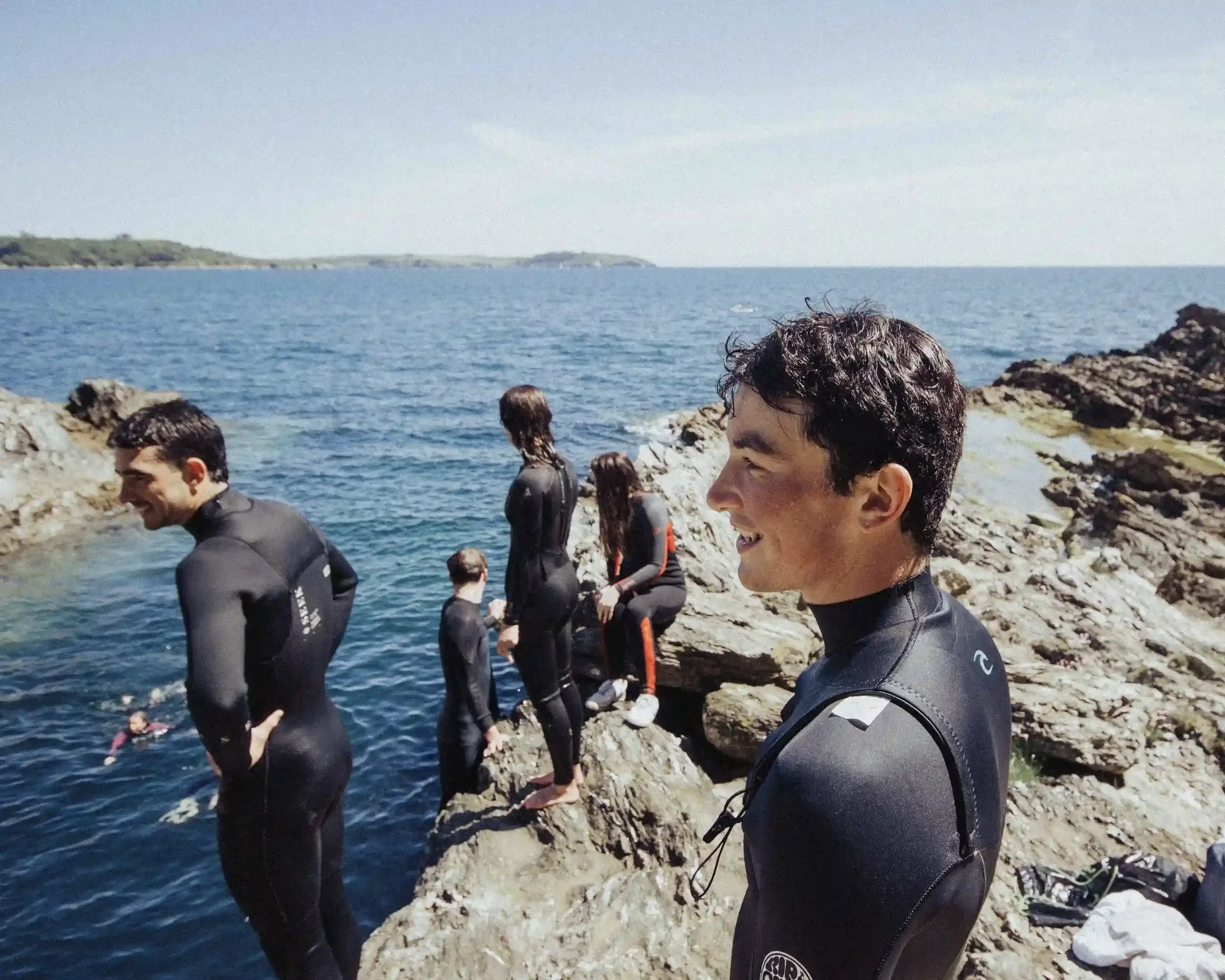 A low-angle profile photograph of singer-songwriter Ned Holland smiling and looking out at the sea. He is in sharp focus, wearing a black Rip Curl wetsuit. In the background, other crew members in wetsuits stand on rugged grey rocks under a clear blu