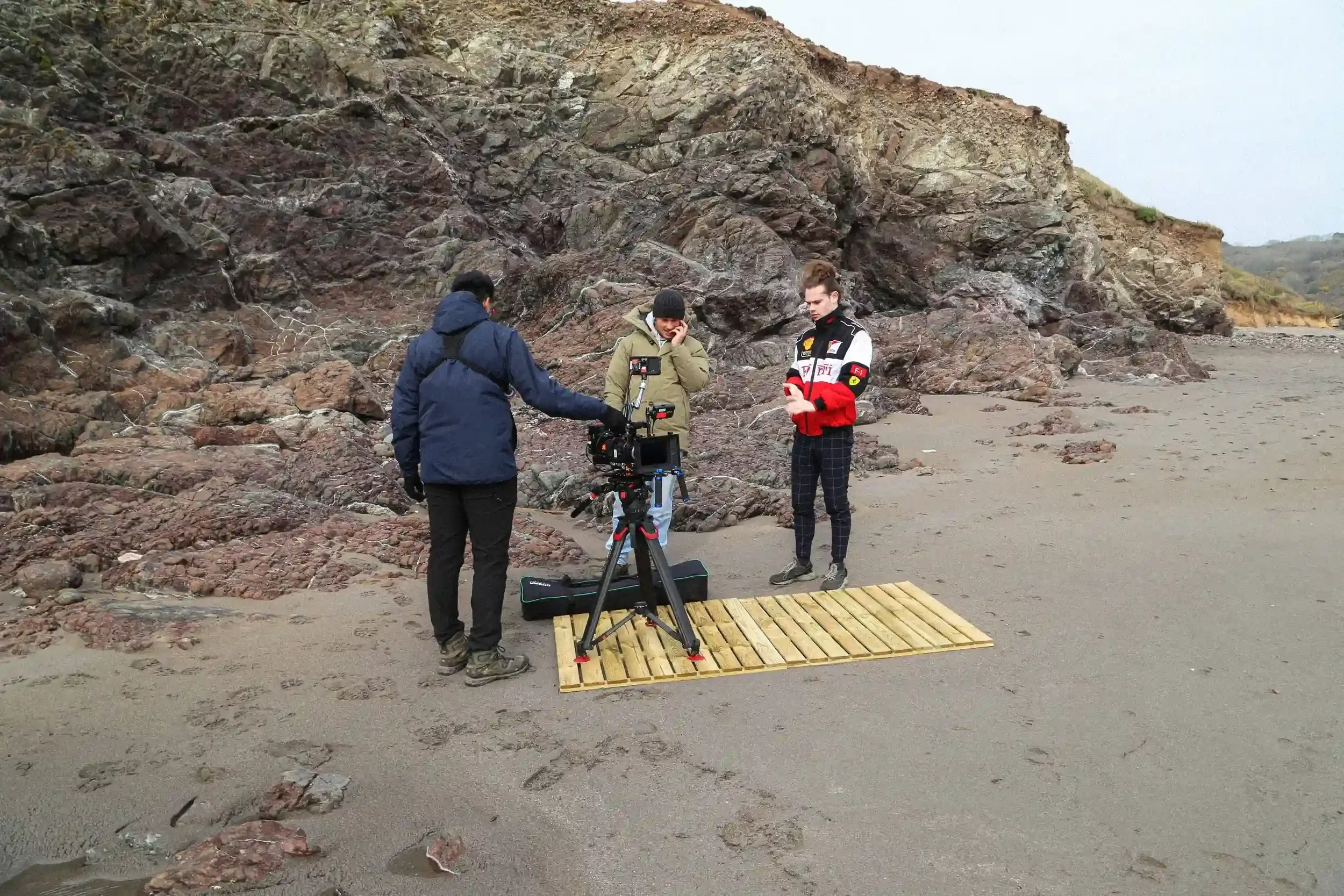 A behind-the-scenes colour photograph of three film crew members standing on a grey sand beach in front of a jagged, dark rock cliff. A professional camera on a tripod is positioned on a wooden pallet laid on the sand. The crew members wear warm jack