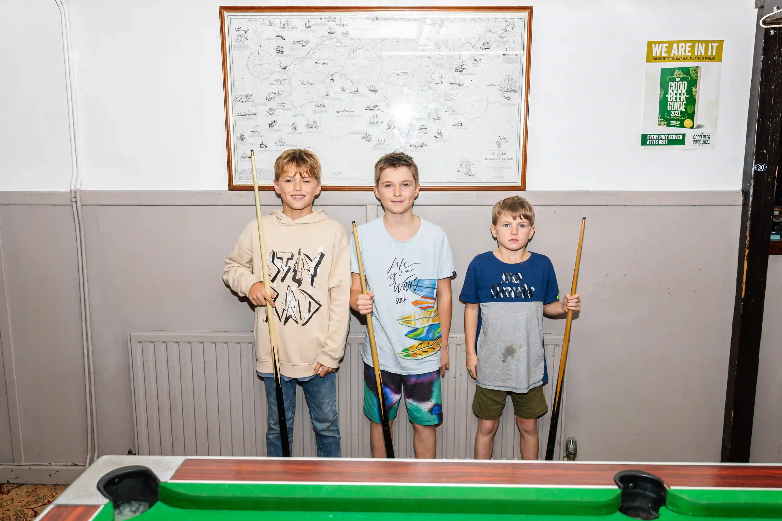 Three young brothers stand side-by-side holding pool cues behind a green-felt pool table in the Seaview Inn, Falmouth. They are positioned against a grey wall featuring a framed nautical map and a 'Good Beer Guide 2021' poster, by Matthew Morgan.