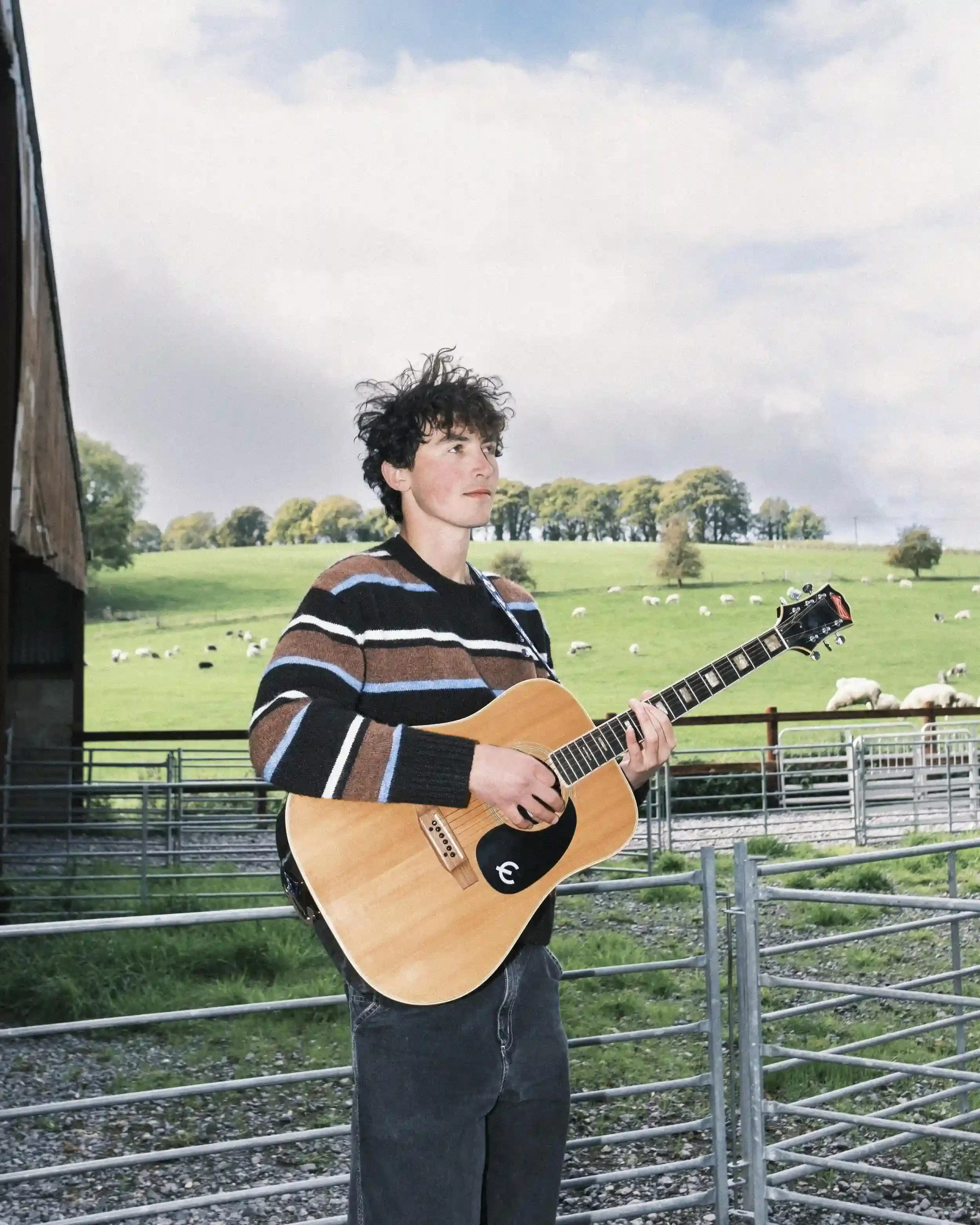 A colour medium-portrait of musician Ned Holland standing in a farmyard in Salisbury. Holland, wearing a dark striped jumper with blue and brown accents, looks off-camera with a thoughtful expression while holding an acoustic guitar. Behind him, a me
