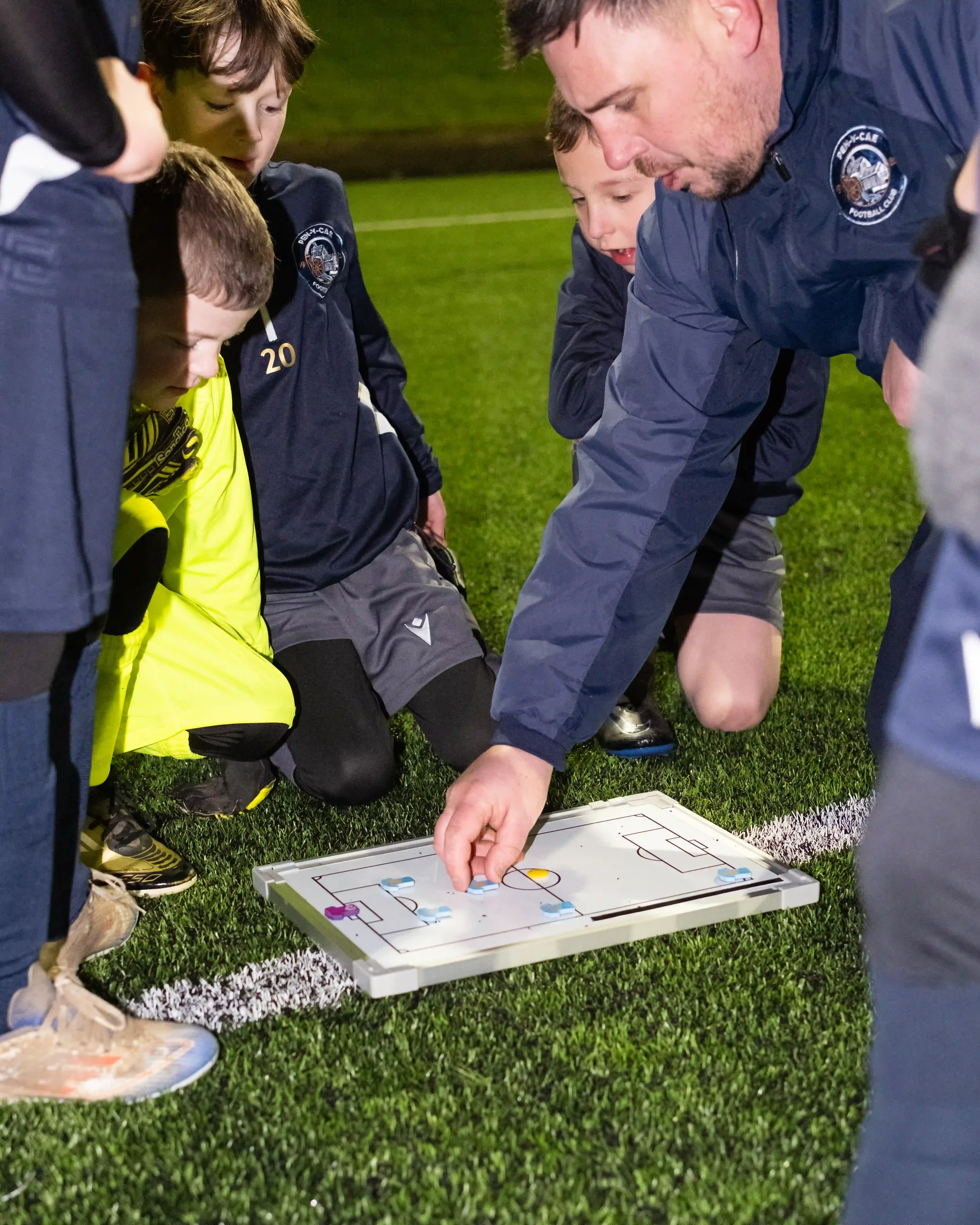 A close-up action shot on a green football pitch at night. A coach in a navy Macron jacket is kneeling, his hand reaching out to move small blue and yellow shirt-shaped magnets on a white tactical board laid on the ground. Several young players in gr