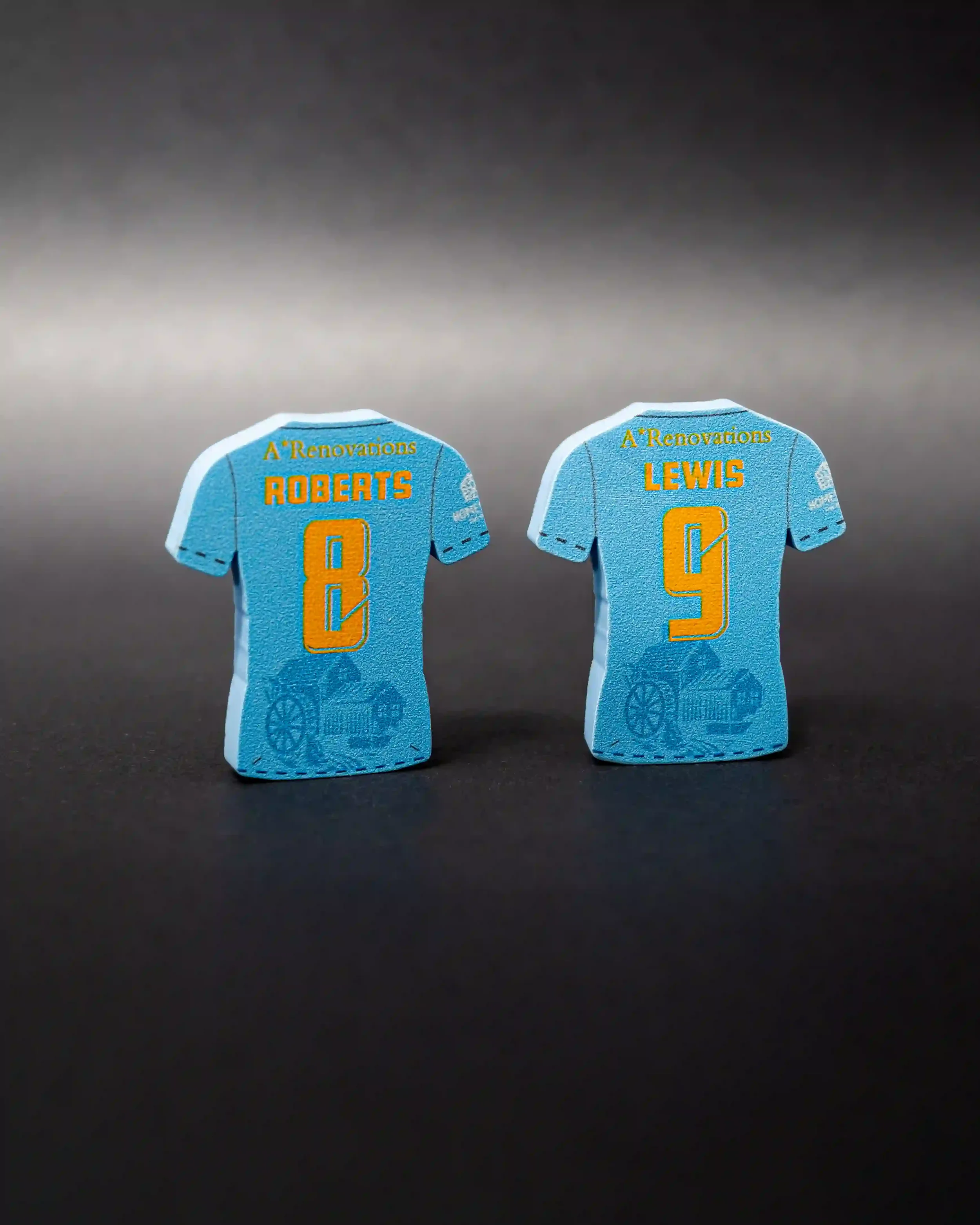 A clean studio product shot of two upright, shirt-shaped magnets standing side-by-side on a dark grey surface. The magnets are a light blue color and show the back of a Pen-Y-Cae FC jersey. The left magnet is personalized with 'ROBERTS 8' and the rig