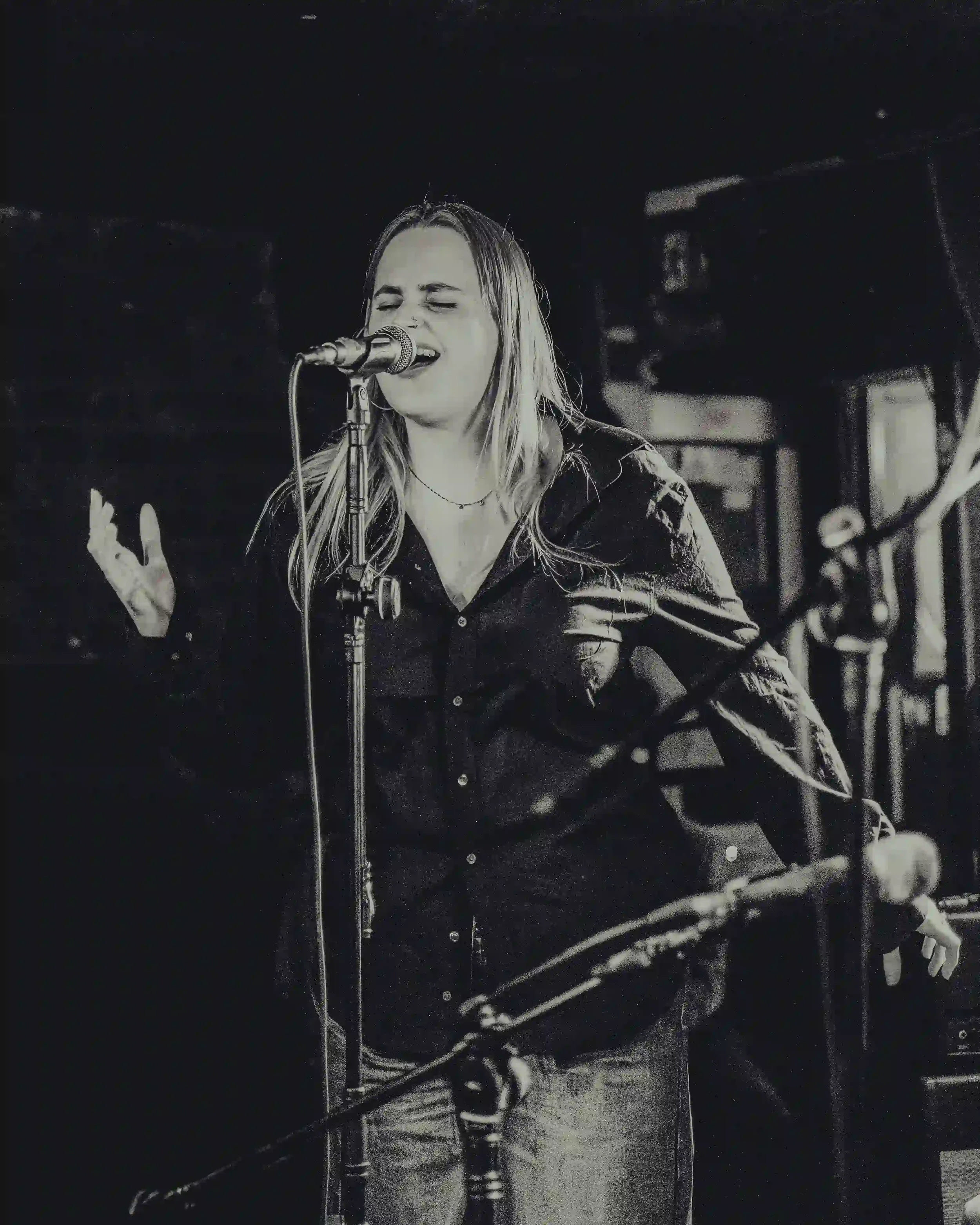 A soulful black-and-white photograph of a female singer performing live on stage at The Troubadour, London. She is captured from the waist up, wearing a dark button-down shirt, singing into a microphone with her eyes closed and her left hand raised e