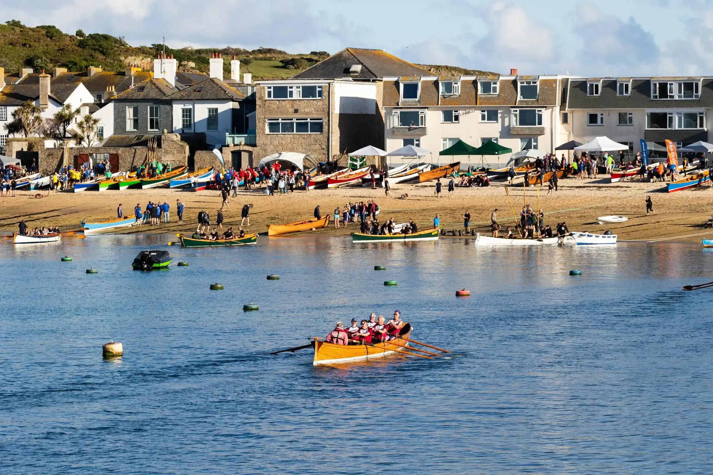 A wide scenic shot of a wooden pilot gig with a crew of seven rowing through calm blue water toward a busy beach in the Isles of Scilly. The shoreline is crowded with people and numerous colorful rowing boats parked on the sand. In the background, wh