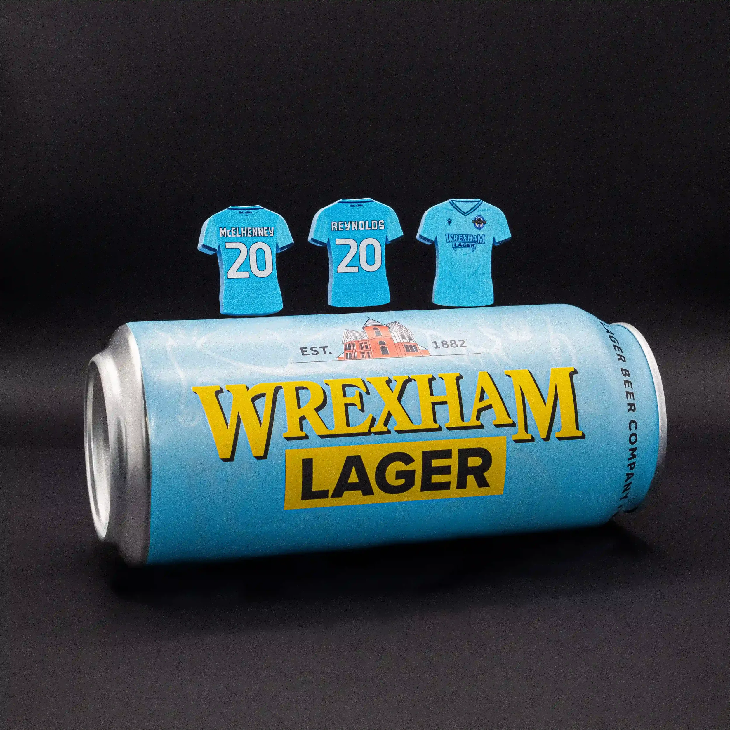 A professional lifestyle product shot of three light blue, shirt-shaped magnets standing behind a horizontal light blue can of Wrexham Lager. The magnets are personalized for Wrexham AFC owners with 'REYNOLDS 20' and 'McELHENNEY 20', alongside a fron