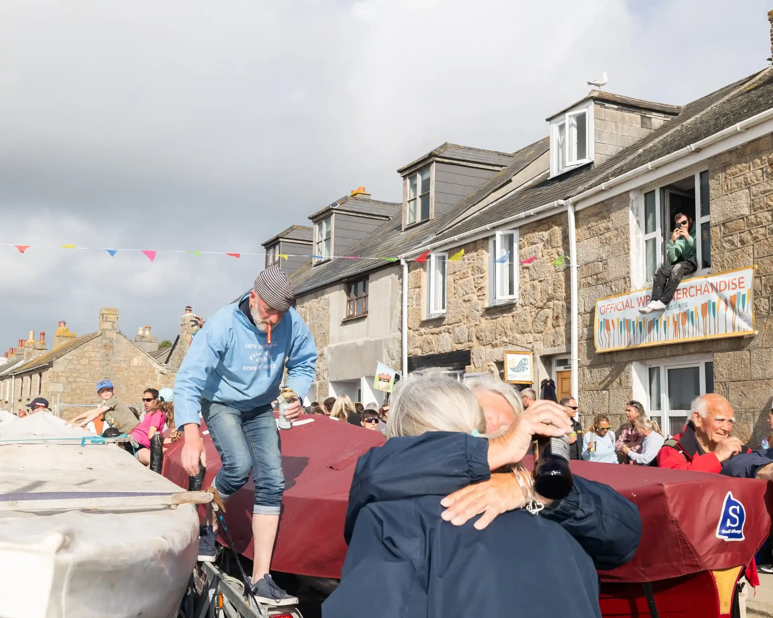 A busy street scene during the World Pilot Gig Championships celebration in the Isles of Scilly. In the foreground, two people are seen from behind in an emotional embrace, one holding a dark glass bottle. To the left, a man in a light blue hoodie an