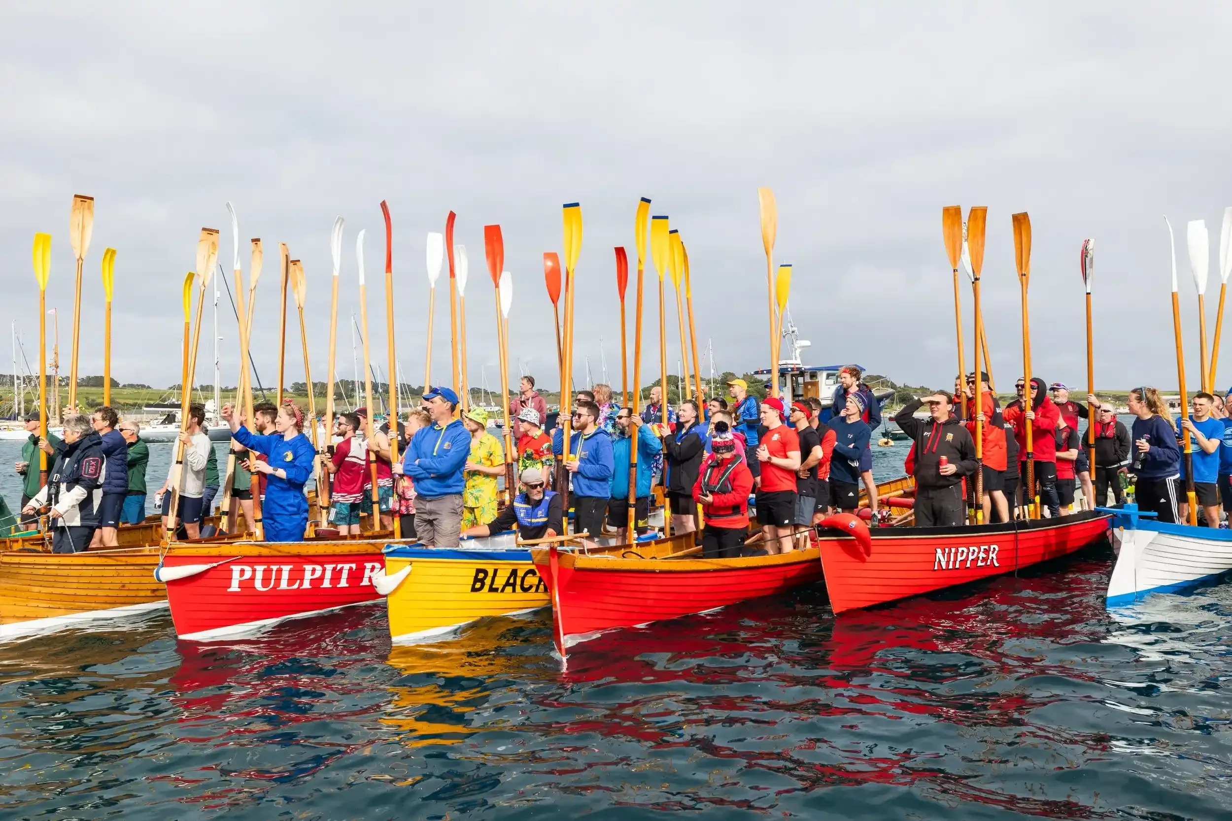 A wide shot of a colorful fleet of wooden pilot gigs on calm blue water during the World Pilot Gig Championships. Each boat is filled with crew members wearing different colored team gear, and they are all holding their long wooden oars vertically to