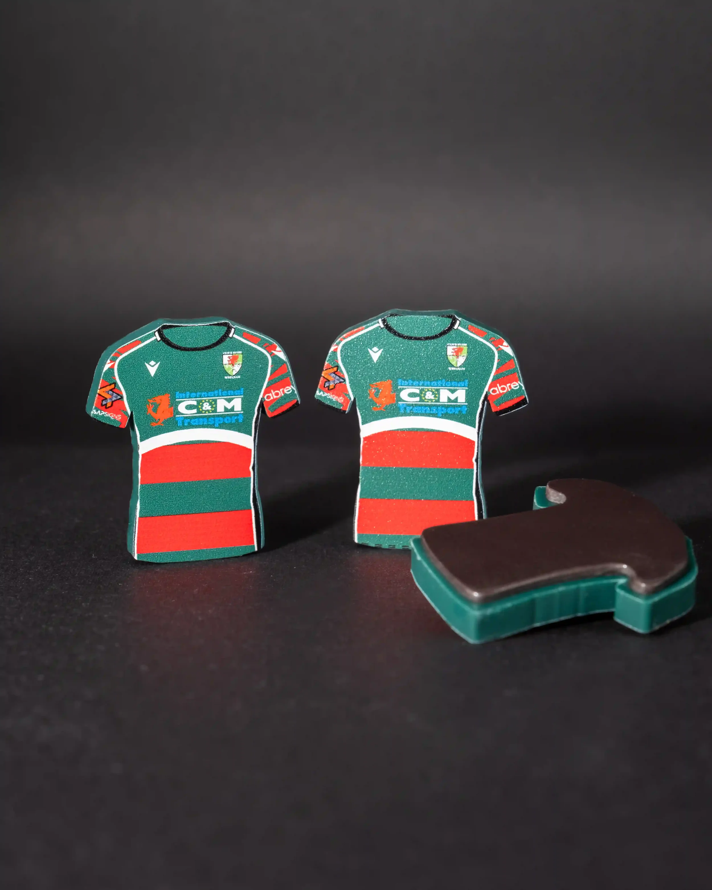 A professional product shot of two upright, shirt-shaped magnets for Wrexham RFC standing on a dark surface. The magnets feature a green upper half with a red and green hooped bottom. They prominently display the club crest, the Macron logo, and 'Int