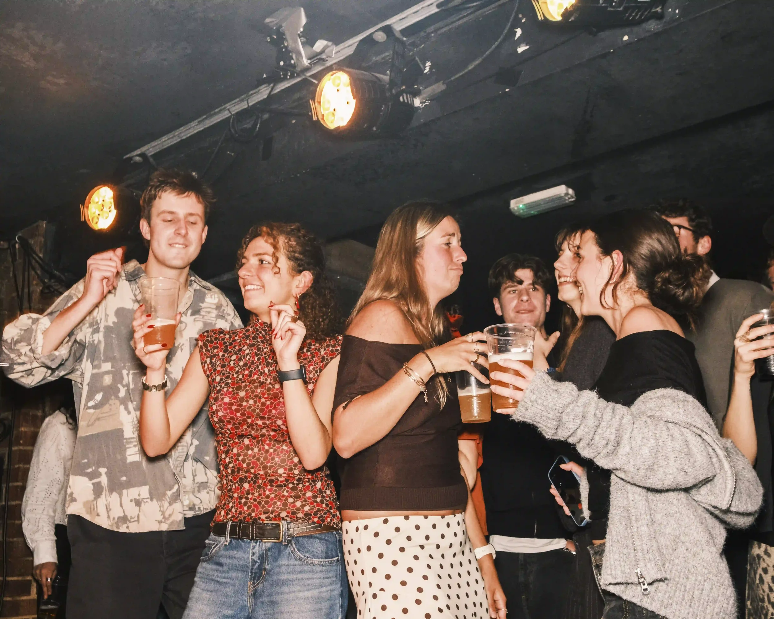 A candid night-time photograph of a young crowd dancing and laughing at The Troubadour in London. Several people are holding plastic cups of beer under warm yellow stage lights that cut through the dark, smoky room. A woman in a polka-dot skirt and a