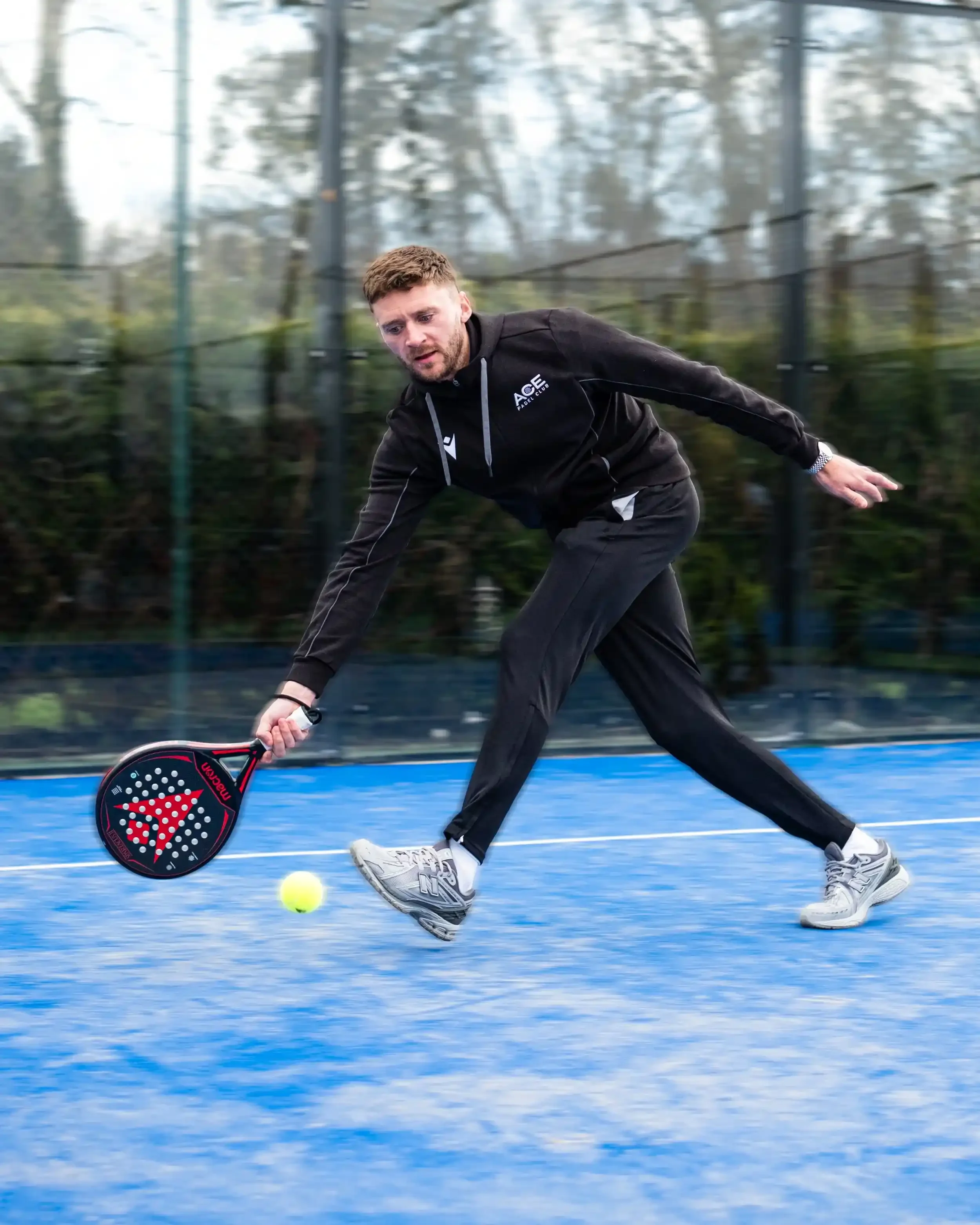 Professional cricketer Joe Clarke in a dynamic action pose on a blue padel court. He is wearing a black Macron tracksuit with the Ace Padel Club logo and is lunging low to return a yellow padel ball with a black and red Macron racket. The background 